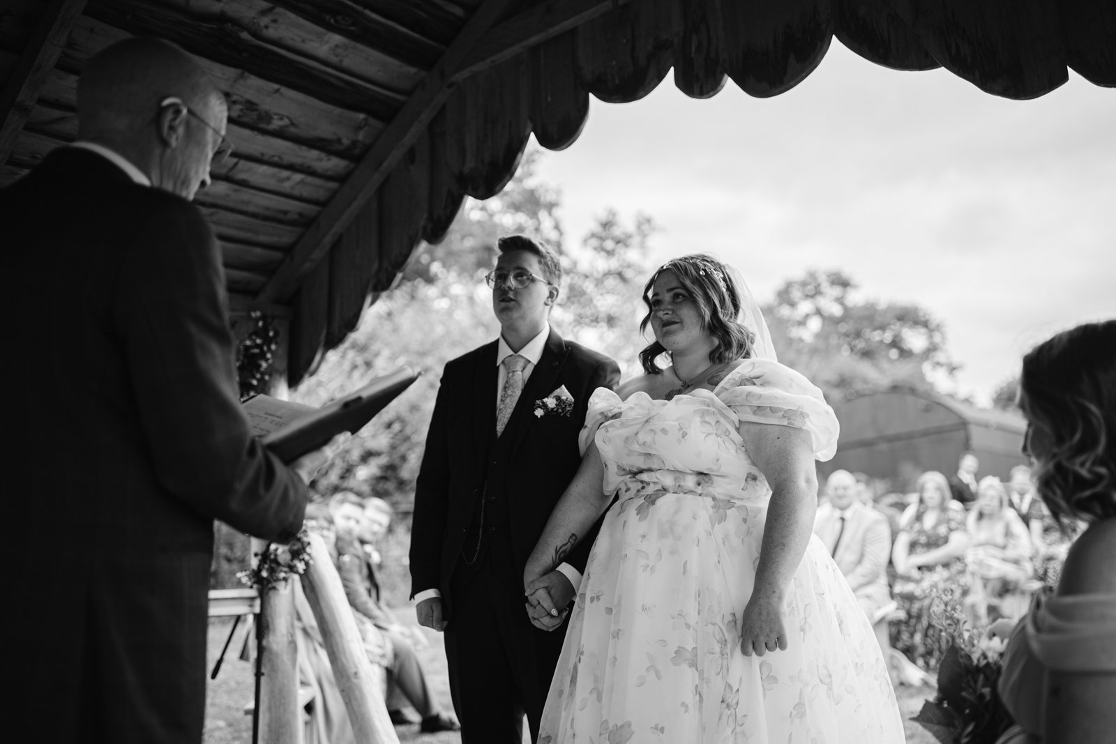 A couple stands holding hands during a rustic farm wedding ceremony in Shrewsbury, facing an officiant, with guests seated in the background at this LGBT+ friendly wedding.