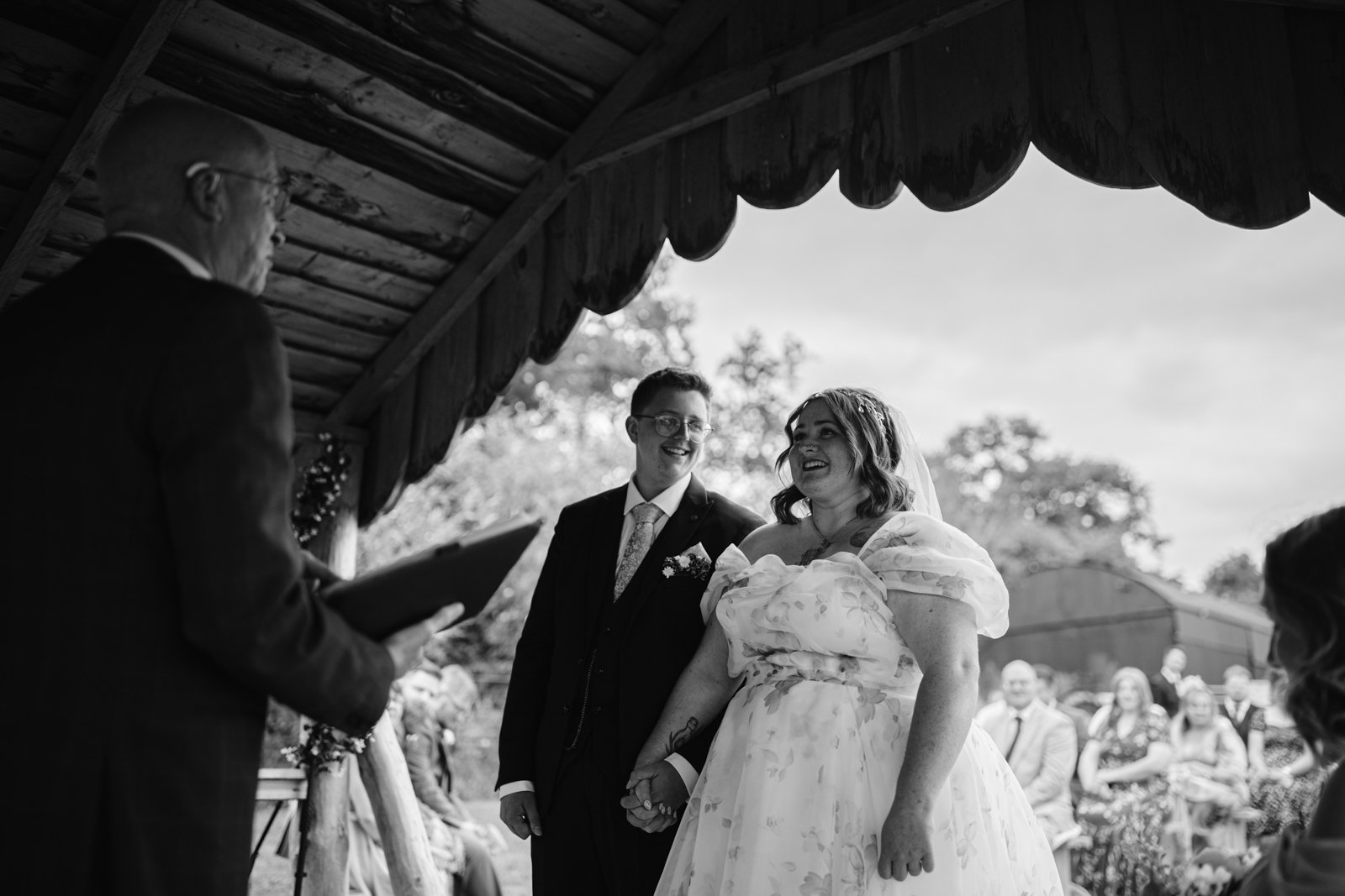 A bride and groom stand holding hands under a wooden pavilion at their rustic farm wedding, facing an officiant during an outdoor ceremony with guests seated in the background.