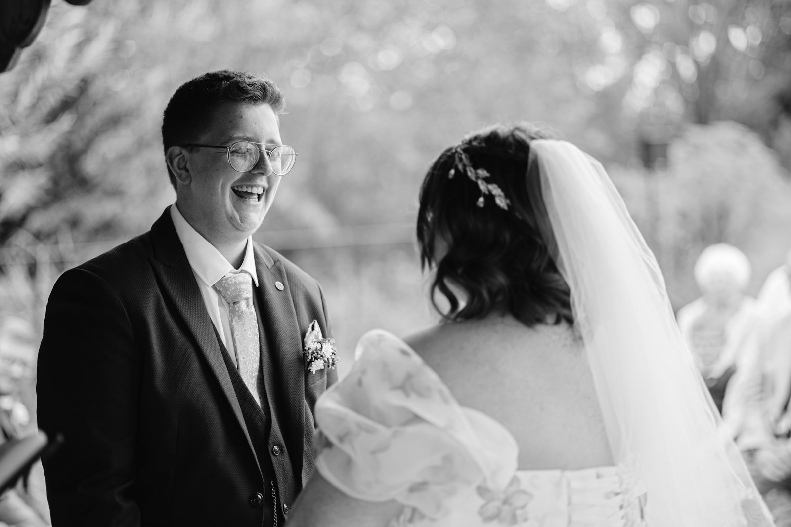 A person in a suit and tie smiles at a person in a wedding dress and veil during an outdoor ceremony at Stanford Farm, capturing the charm of a rustic wedding.