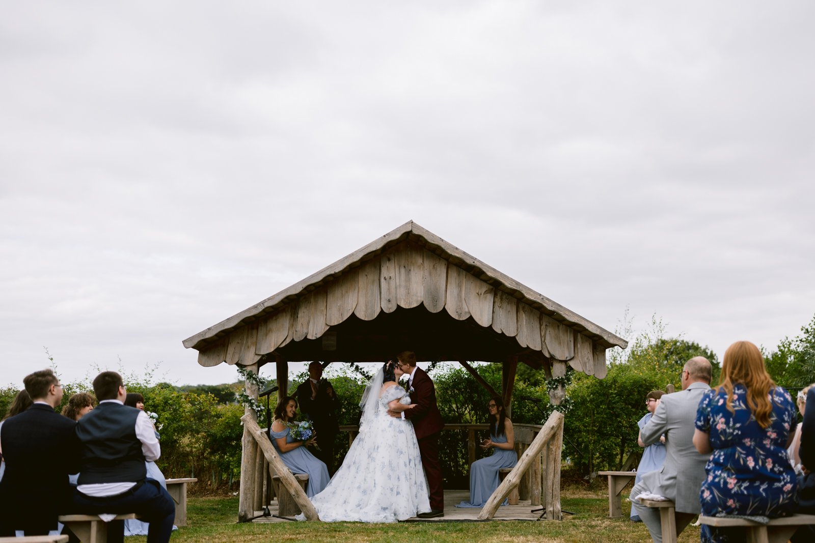 A bride and groom kiss under a rustic wooden gazebo during an outdoor Shrewsbury Wedding ceremony, with guests seated on benches on either side.