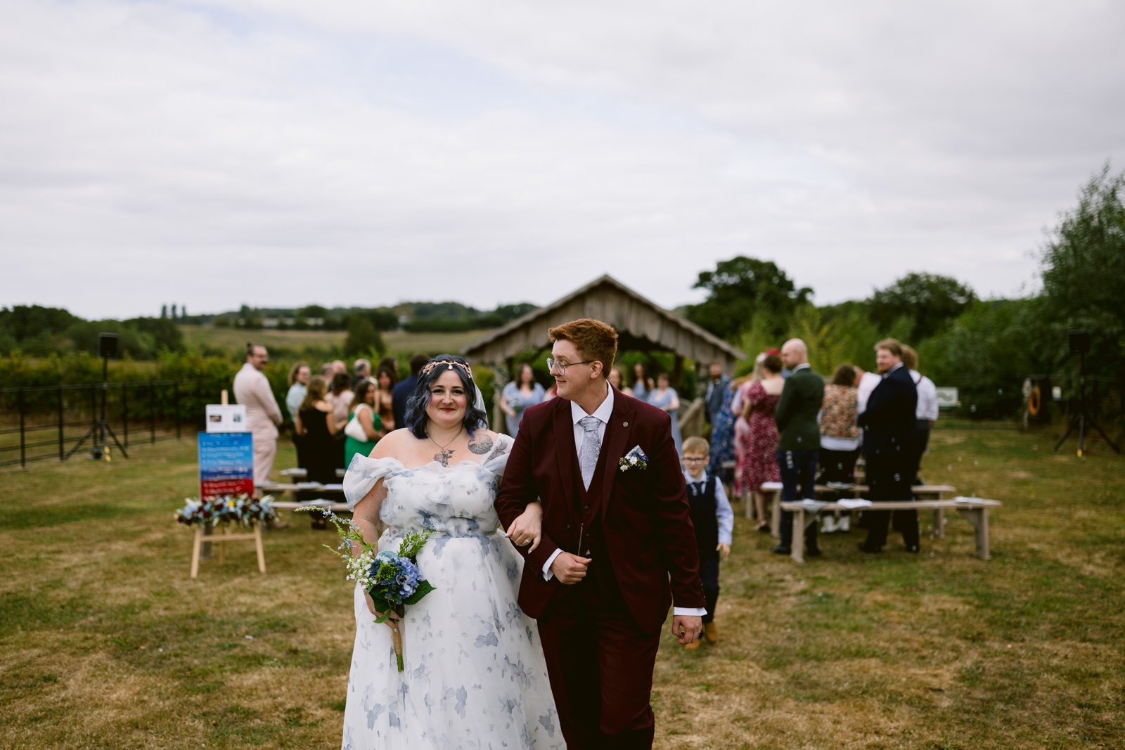 A bride in a white floral dress and a groom in a burgundy suit walk arm in arm outdoors at a rustic farm wedding near Shrewsbury, with guests and a wooden structure visible in the background.