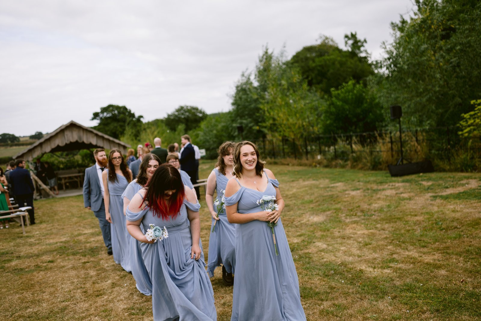 A group of bridesmaids in light blue dresses walk in a line outdoors on a grassy area at a rustic farm wedding, holding bouquets, with guests and trees in the background.