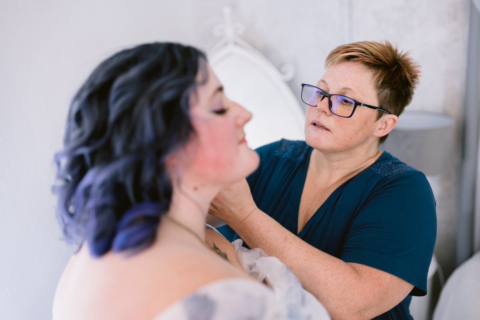 A woman with short hair and glasses helps another woman with dark wavy hair adjust her necklace in a softly lit room, capturing a tender moment at an LGBT friendly wedding.
