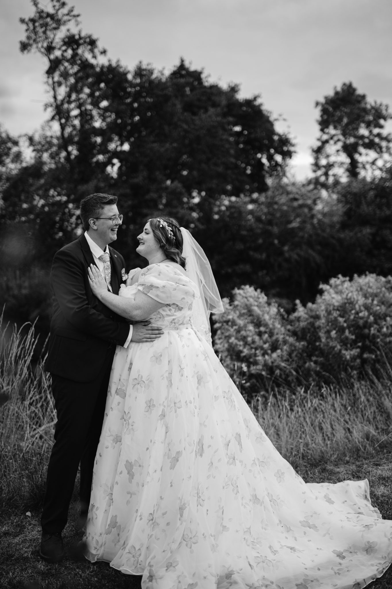 A couple in formal wedding attire stands outdoors, smiling at each other amid trees and bushes at a rustic Stanford Farm Wedding. The black and white image captures a joyful, LGBT+ friendly celebration in Shrewsbury.