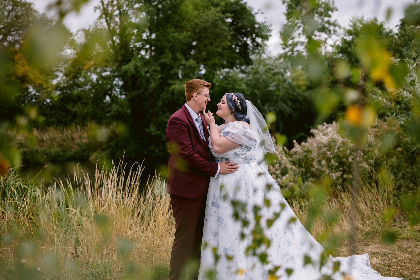 A bride and groom stand outdoors in a grassy area, gazing at each other and holding hands, surrounded by green foliage and trees at a rustic farm wedding.