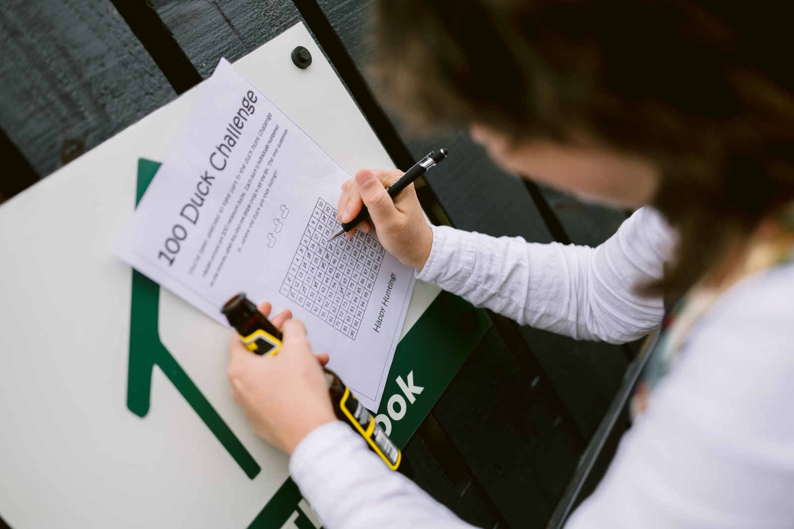 A person marks a word search puzzle titled "100 Duck Challenge" on a piece of paper while holding a yellow device, next to a sign with a green arrow—perfect inspiration for an LGBT+ friendly wedding or rustic farm wedding in Shrewsbury.
