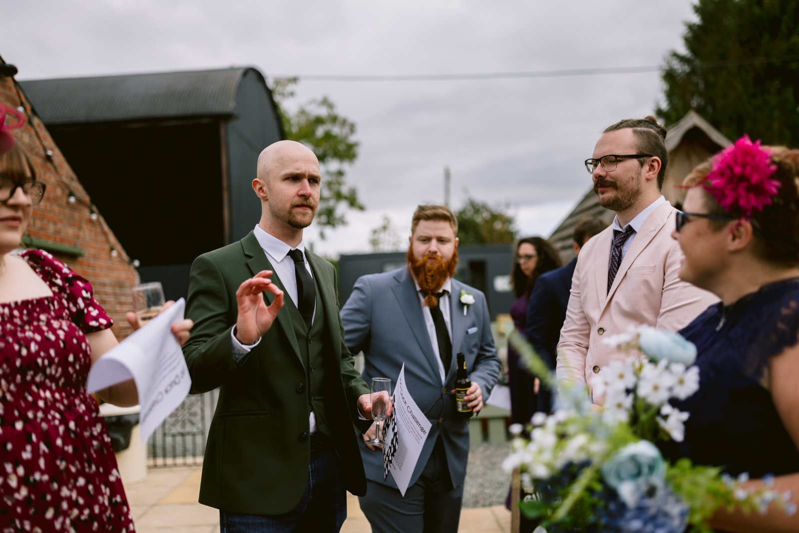 A group of people in formal attire stand outside at a rustic Stanford Farm wedding near Shrewsbury, conversing warmly. One man holds a glass and gestures while others listen. Flowers and papers are visible in the foreground.