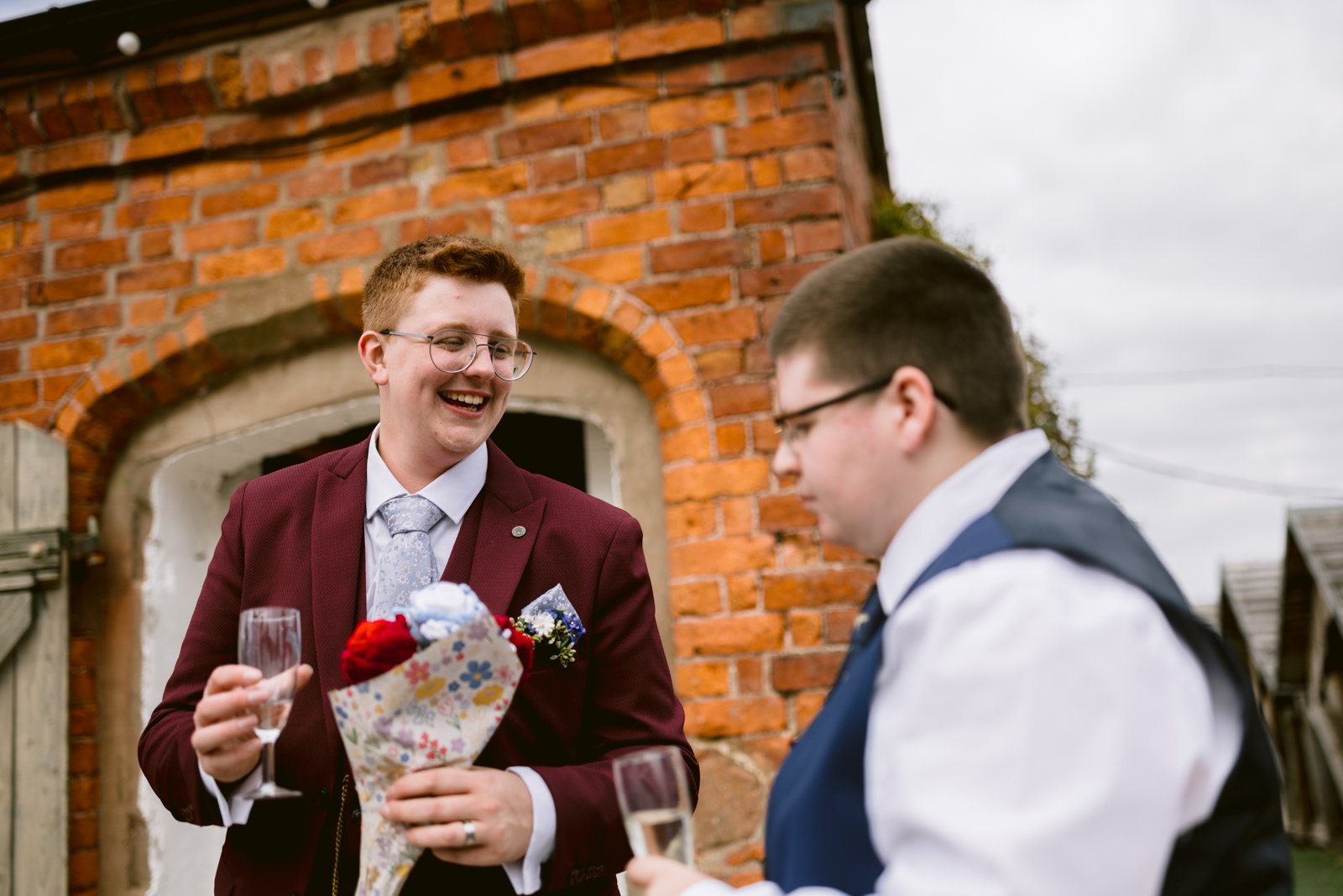 Two people in formal attire stand outside, one holding a bouquet and a champagne glass, both smiling and engaged in conversation in front of a brick building at a rustic farm wedding.