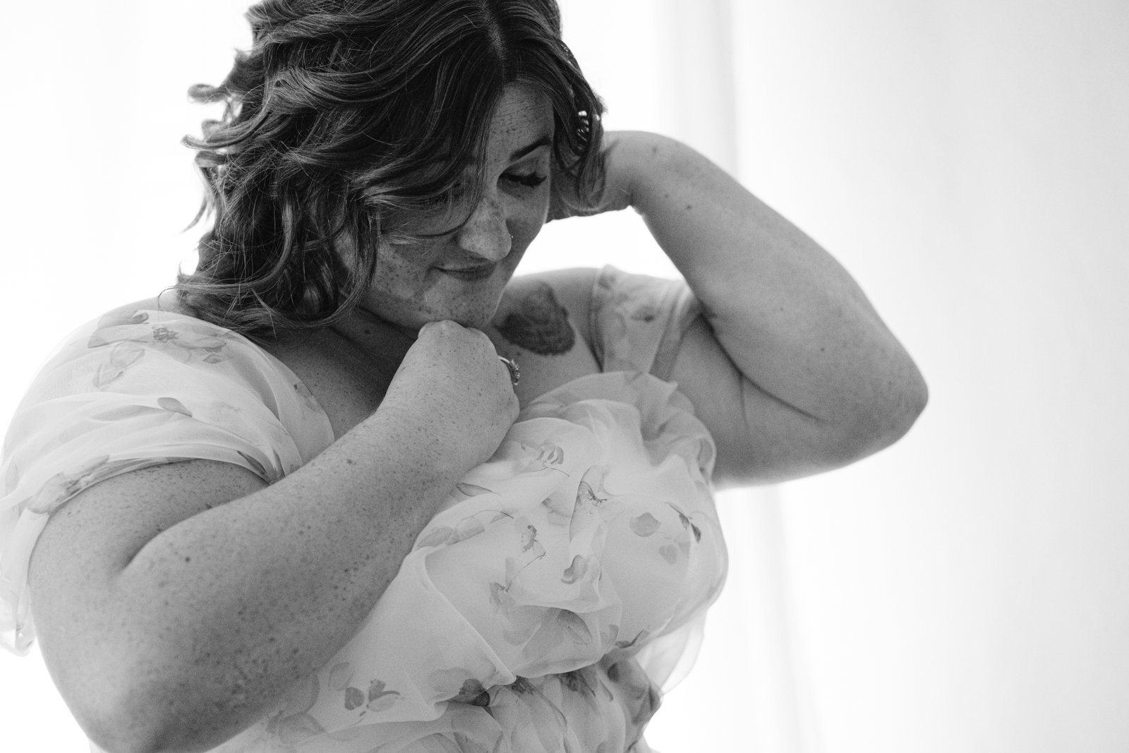 A woman in a floral dress adjusts the fabric near her shoulder, looking down slightly. The black and white photo captures a quiet moment at a rustic Stanford Farm wedding near Shrewsbury.