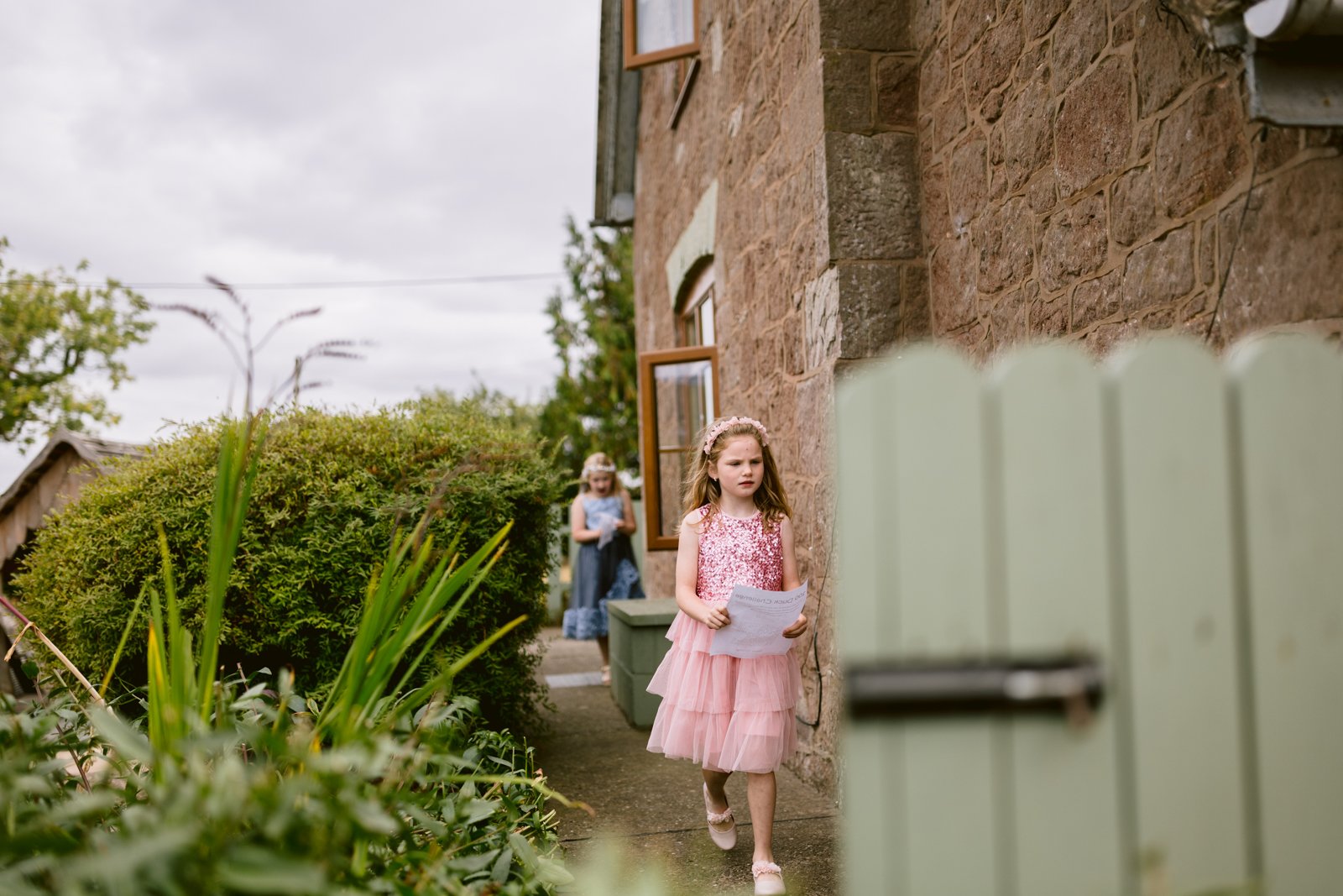 Two young girls walk outside a stone house at a rustic farm wedding; the girl in front wears a pink dress and holds paper, while another follows in the background.