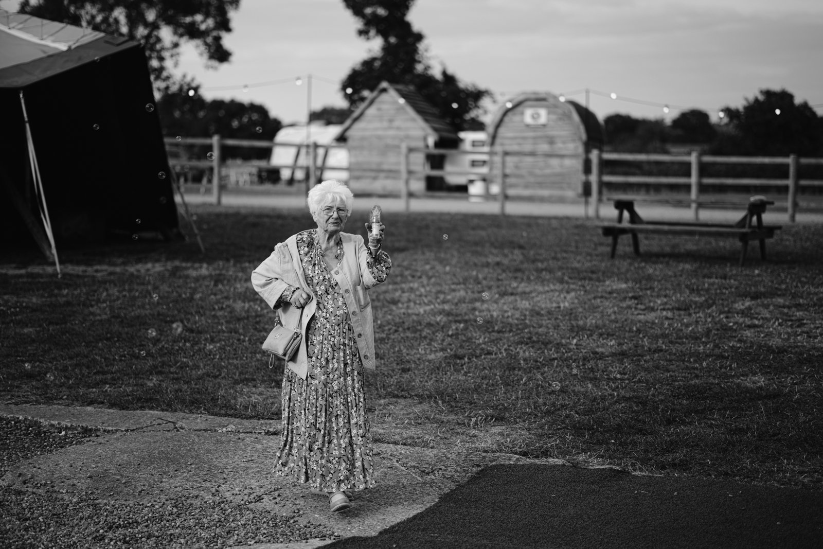 An elderly woman in a floral dress and coat stands on grass at a rustic farm wedding, holding an ice cream cone and a handbag. Wooden huts and a fence complete the charming countryside scene in the background.