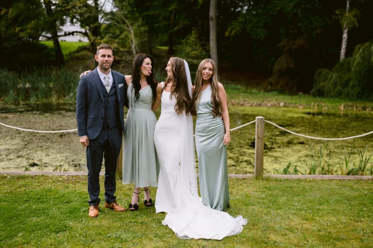 Four people stand side by side outdoors in front of a pond for a classic Wedding Group Photo, with three women in light green dresses and one man in a blue suit, all dressed in formal attire.
