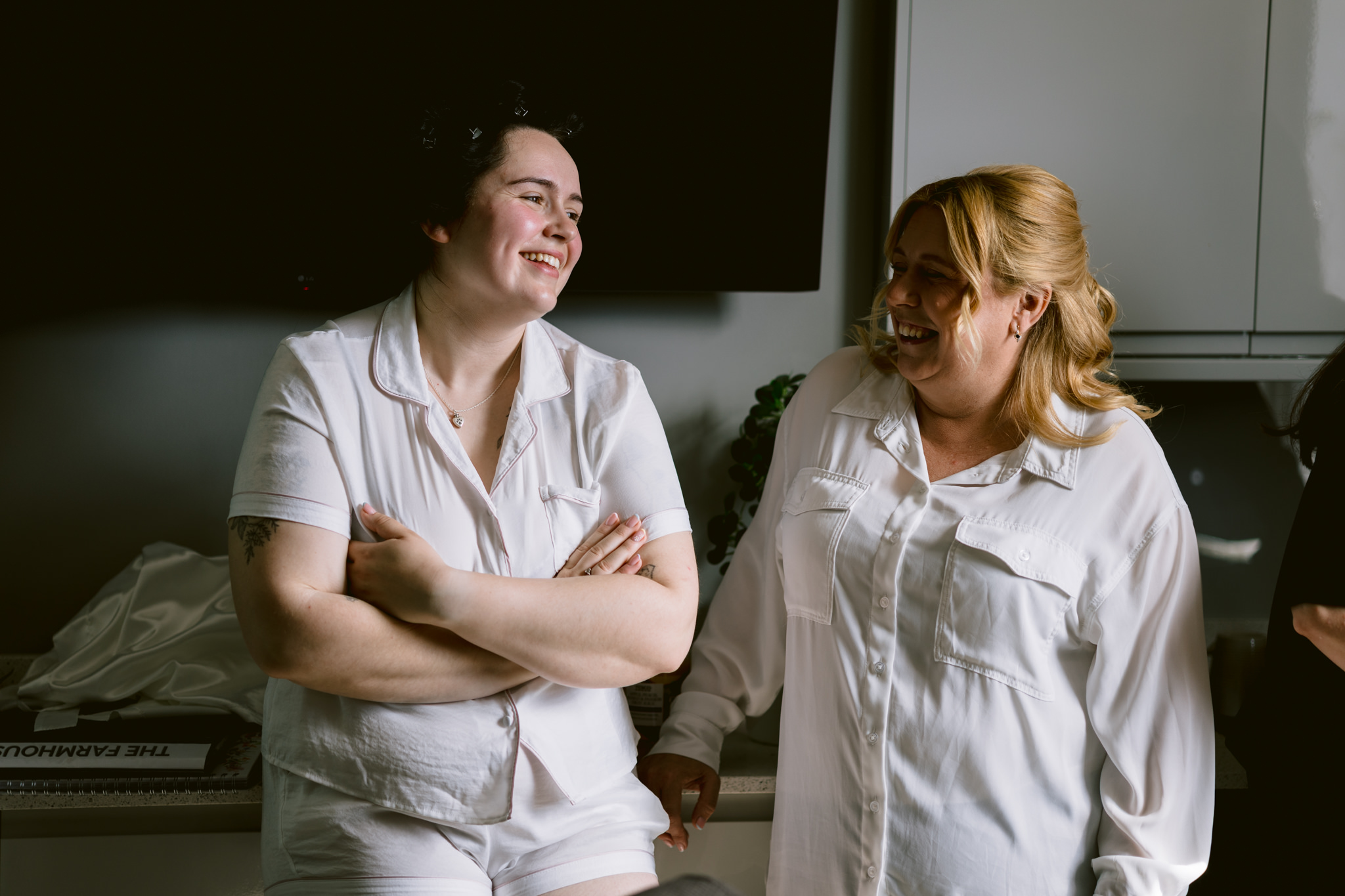Two people wearing white pyjamas stand indoors, smiling and talking to each other, with a kitchen background—capturing a candid moment reminiscent of relaxed wedding photography at an Essendon Country Club wedding.