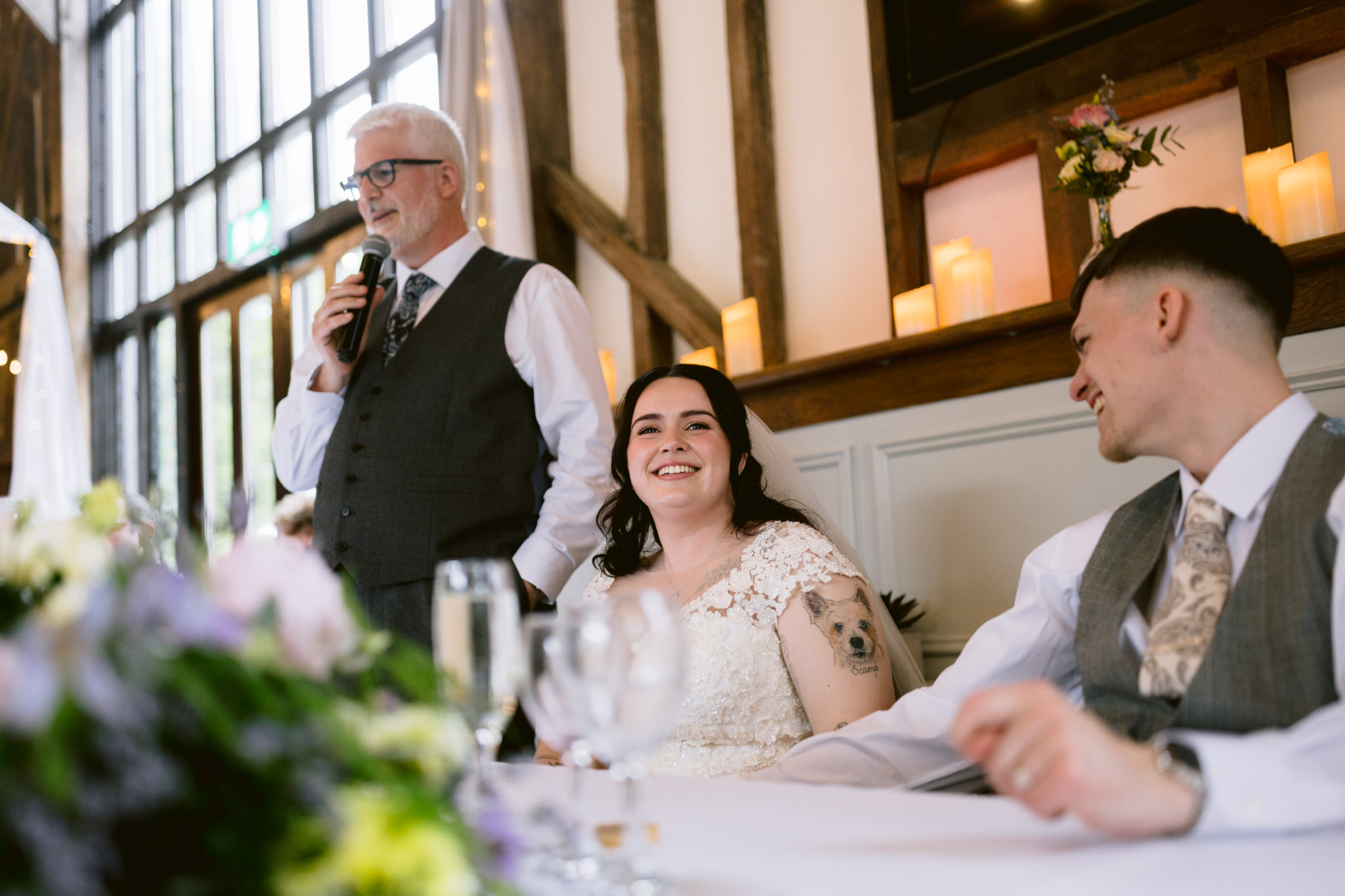 A man gives a heartfelt speech with a microphone whilst the bride smiles and the groom looks at her, seated at a decorated table in a bright, rustic venue—perfect for Barn Wedding Hertfordshire memories.