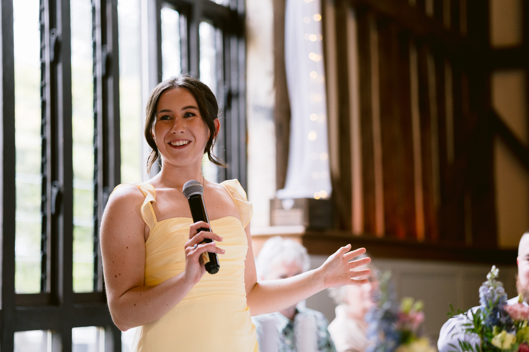 A woman in a yellow dress speaks into a microphone at an indoor event, standing near large windows with guests seated in the background—a candid moment captured by Essendon Country Club Wedding Photography.