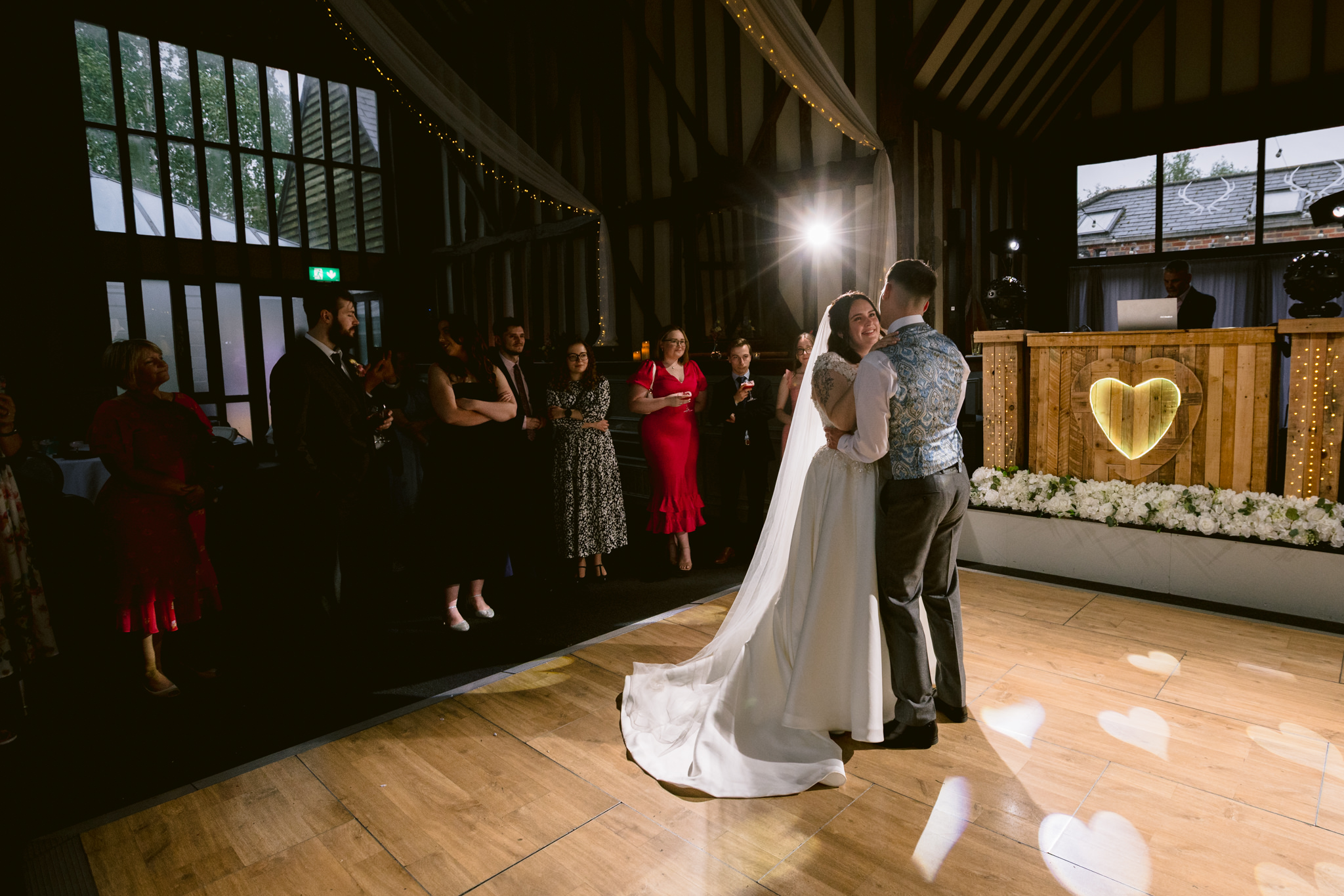 A bride and groom share their first dance on a wooden floor at a charming barn wedding in Hertfordshire, as guests watch; fairy lights and a heart decoration create the perfect backdrop for wedding photography.