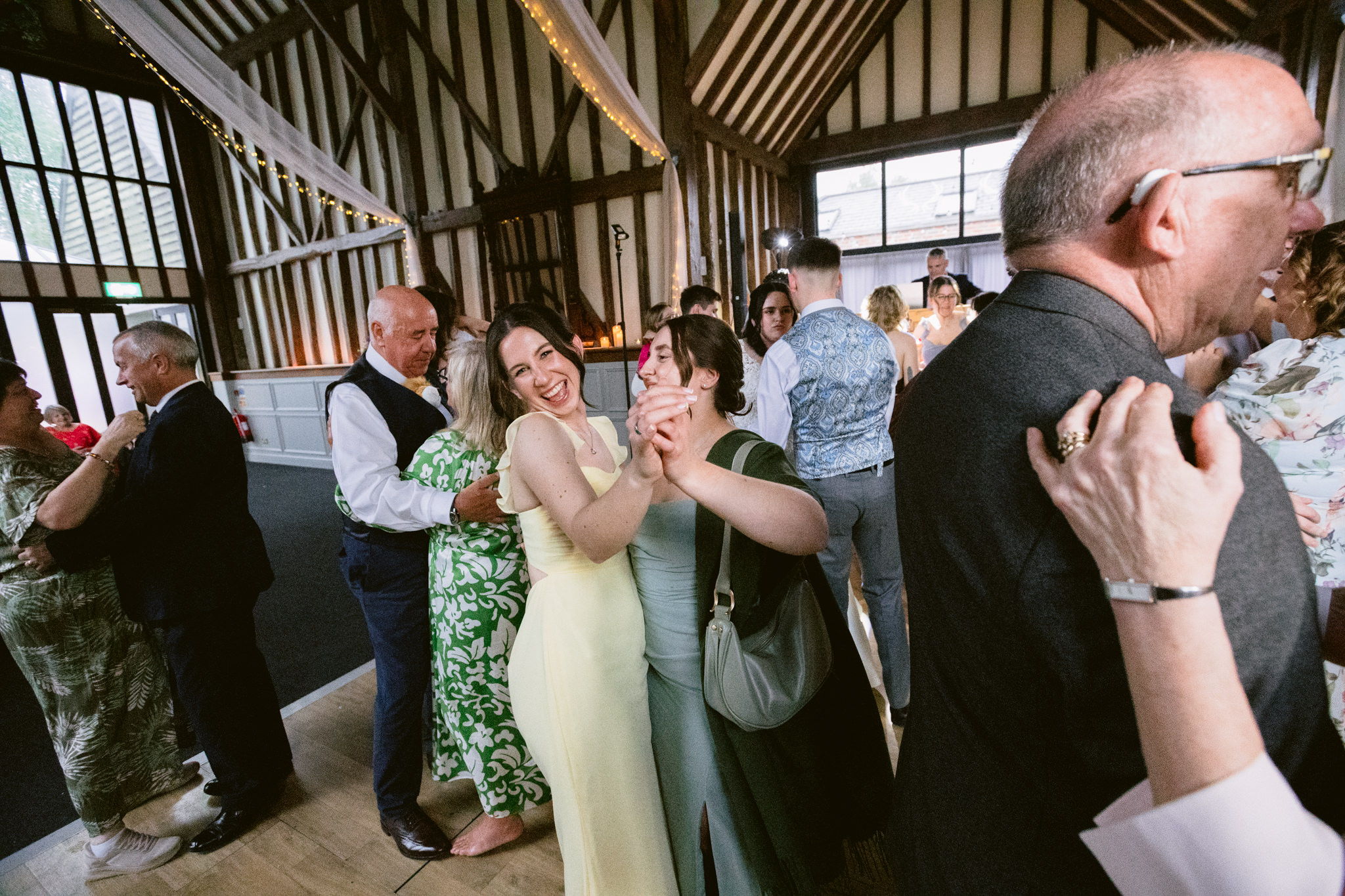 Couples dance in pairs inside a rustic wooden hall with high ceilings and exposed beams at Essendon Country Club, Hertfordshire. Several couples are smiling and enjoying the lively atmosphere, perfect for wedding photography.