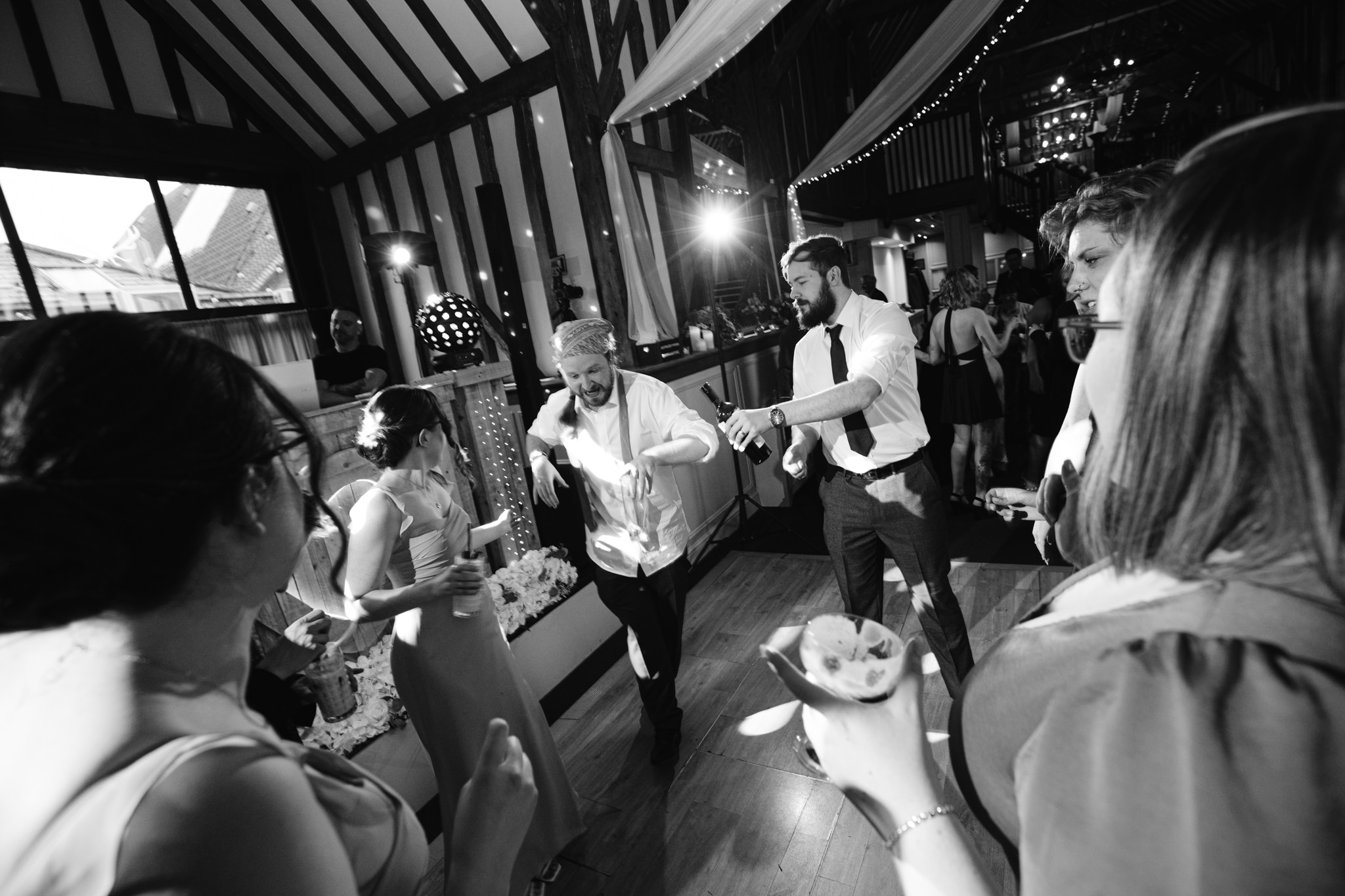 A group of people dressed in formal attire dance and socialise at an indoor Barn Wedding Hertfordshire, with wooden beams and fairy lights overhead.