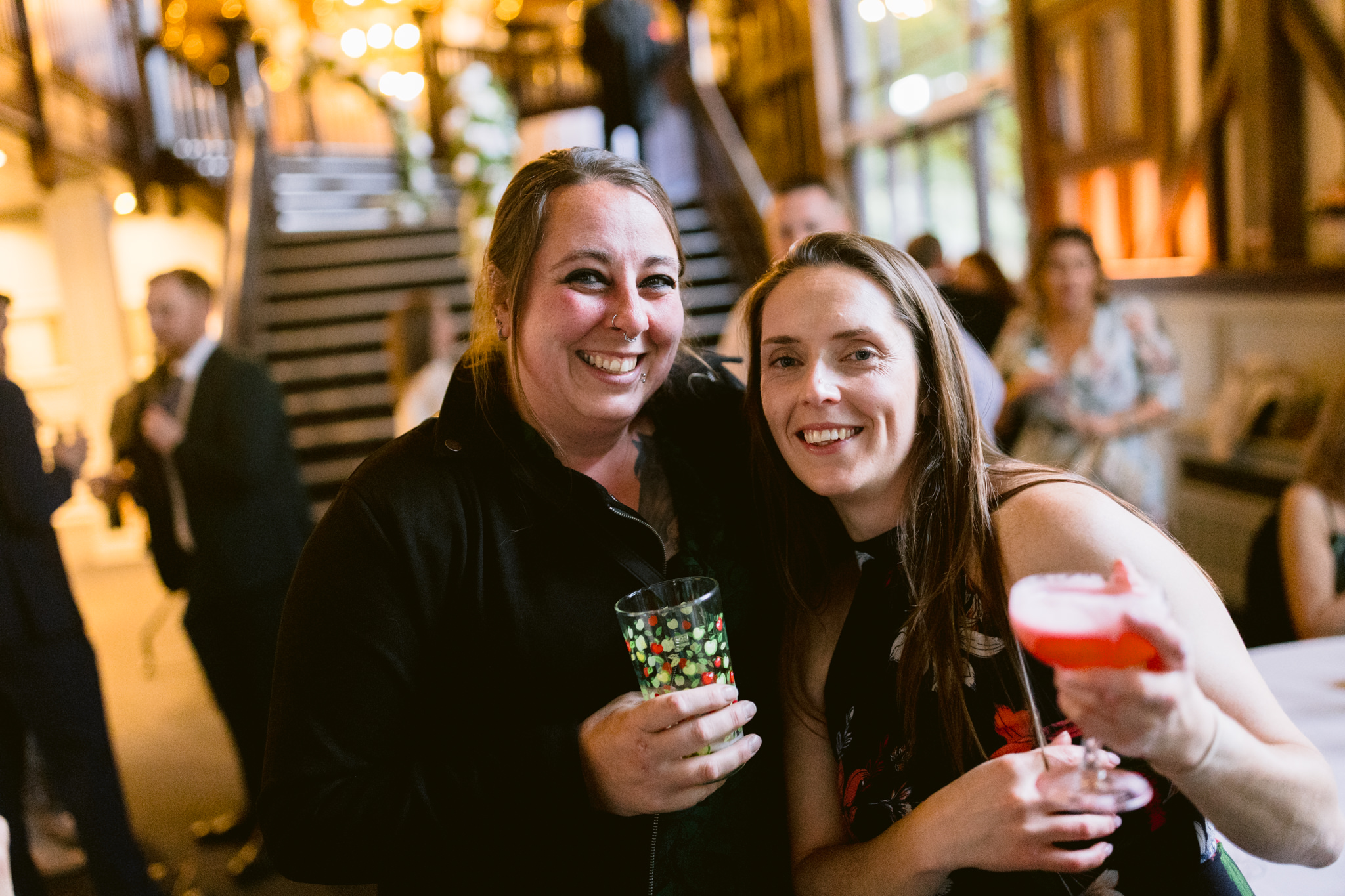 Two women smiling and holding drinks at an indoor Essendon Country Club wedding, with other guests and a staircase visible in the background.