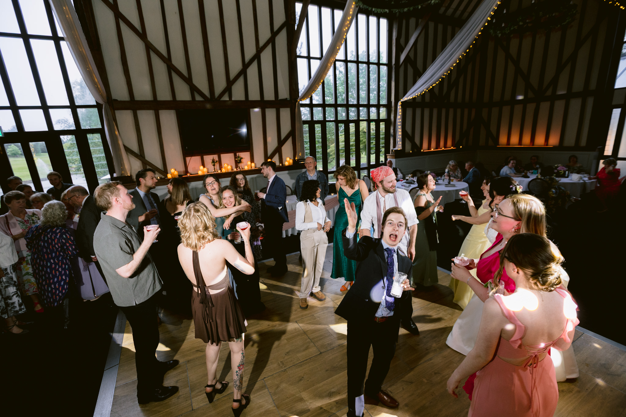 People dancing and socialising at a wedding reception in a barn-style venue with wooden beams and large windows, beautifully captured by wedding photography at the stunning Essendon Country Club in Hertfordshire.
