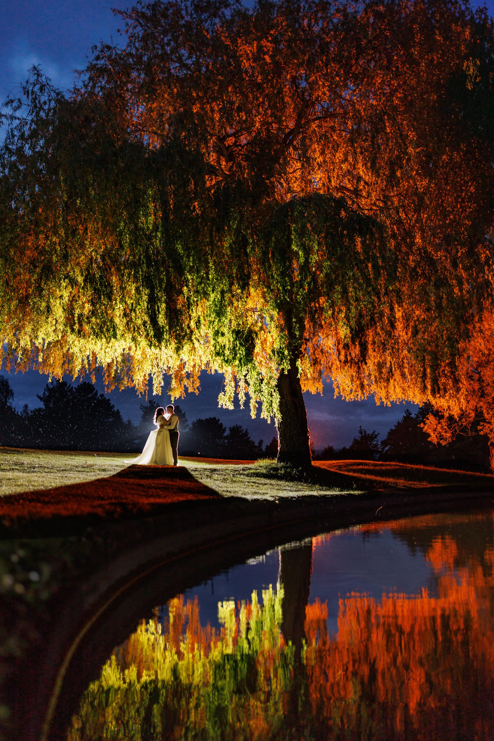 A couple stands under a large, illuminated tree at dusk, their reflection shimmering in a calm pond nearby—a magical moment of Essendon Country Club Wedding elegance.