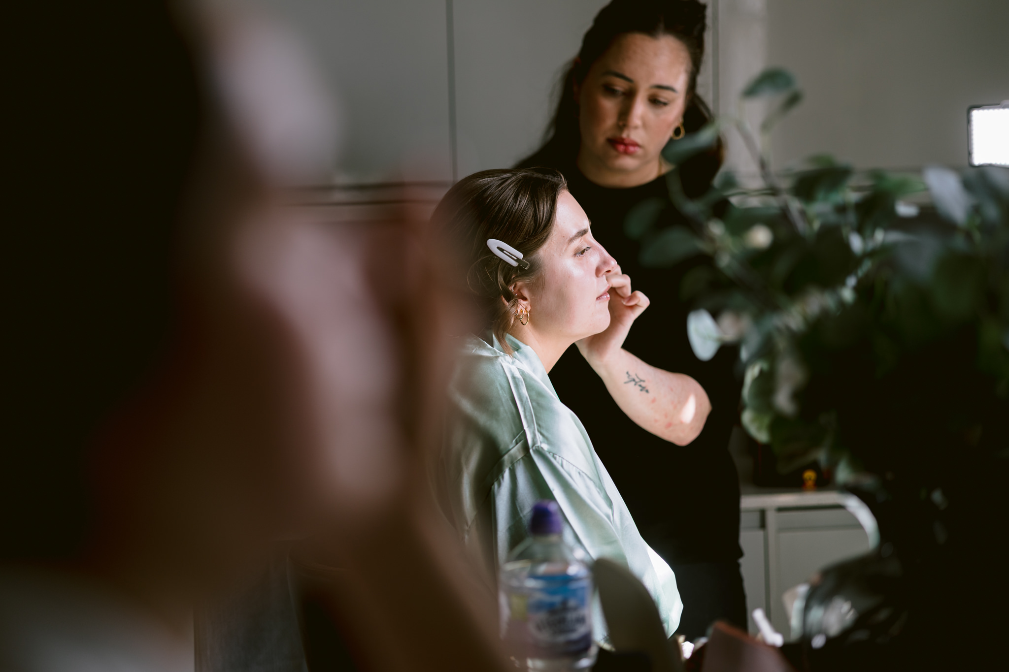 A woman applies make-up to another woman, seated with a hair clip, in a softly lit indoor setting with plants in the foreground—a serene moment captured by Hertfordshire Wedding Photography.