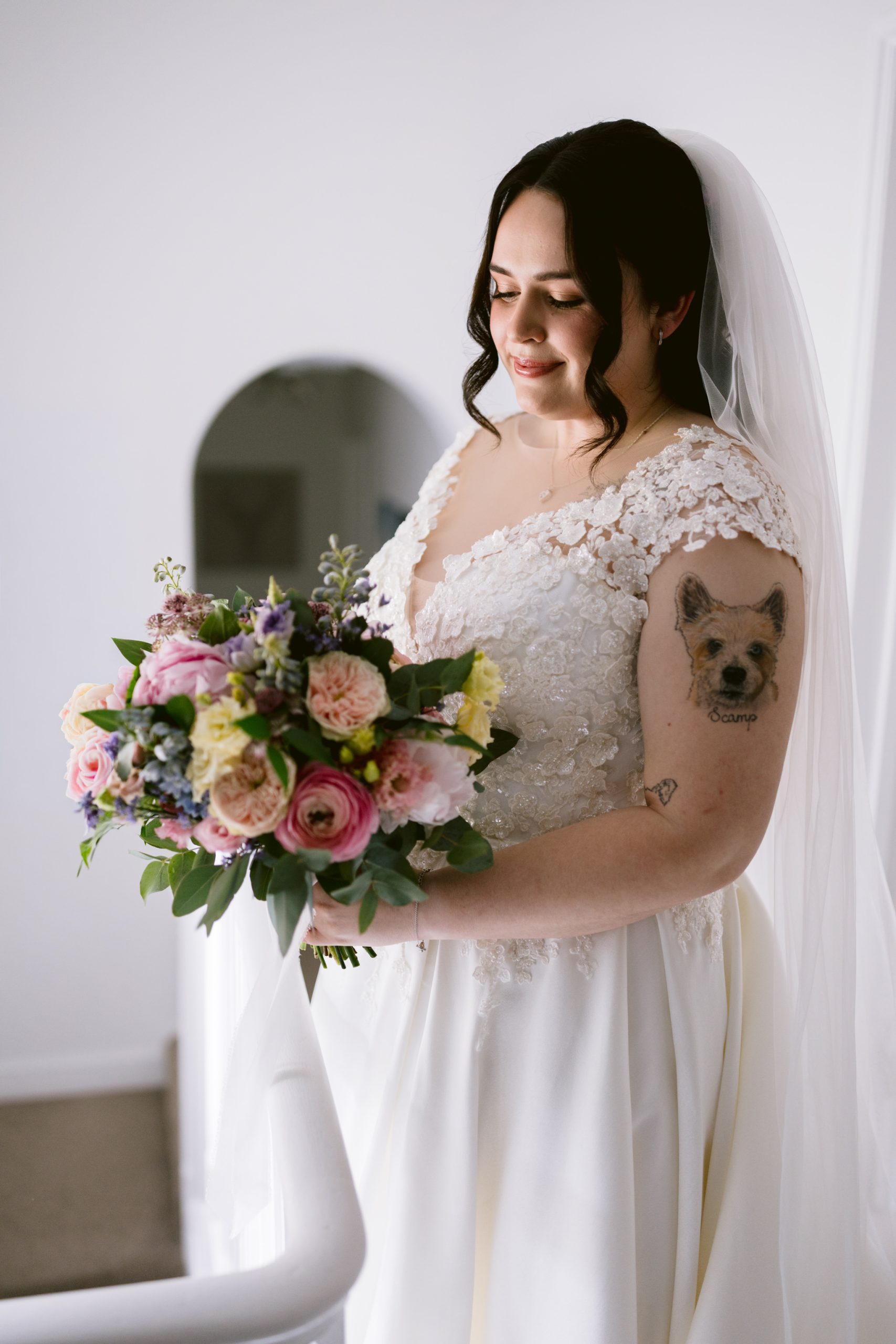 A bride in a white lace wedding dress and veil holds a bouquet of pink and purple flowers; her dog tattoo peeps out on her upper arm, capturing the joy of a Barn Wedding Hertfordshire.