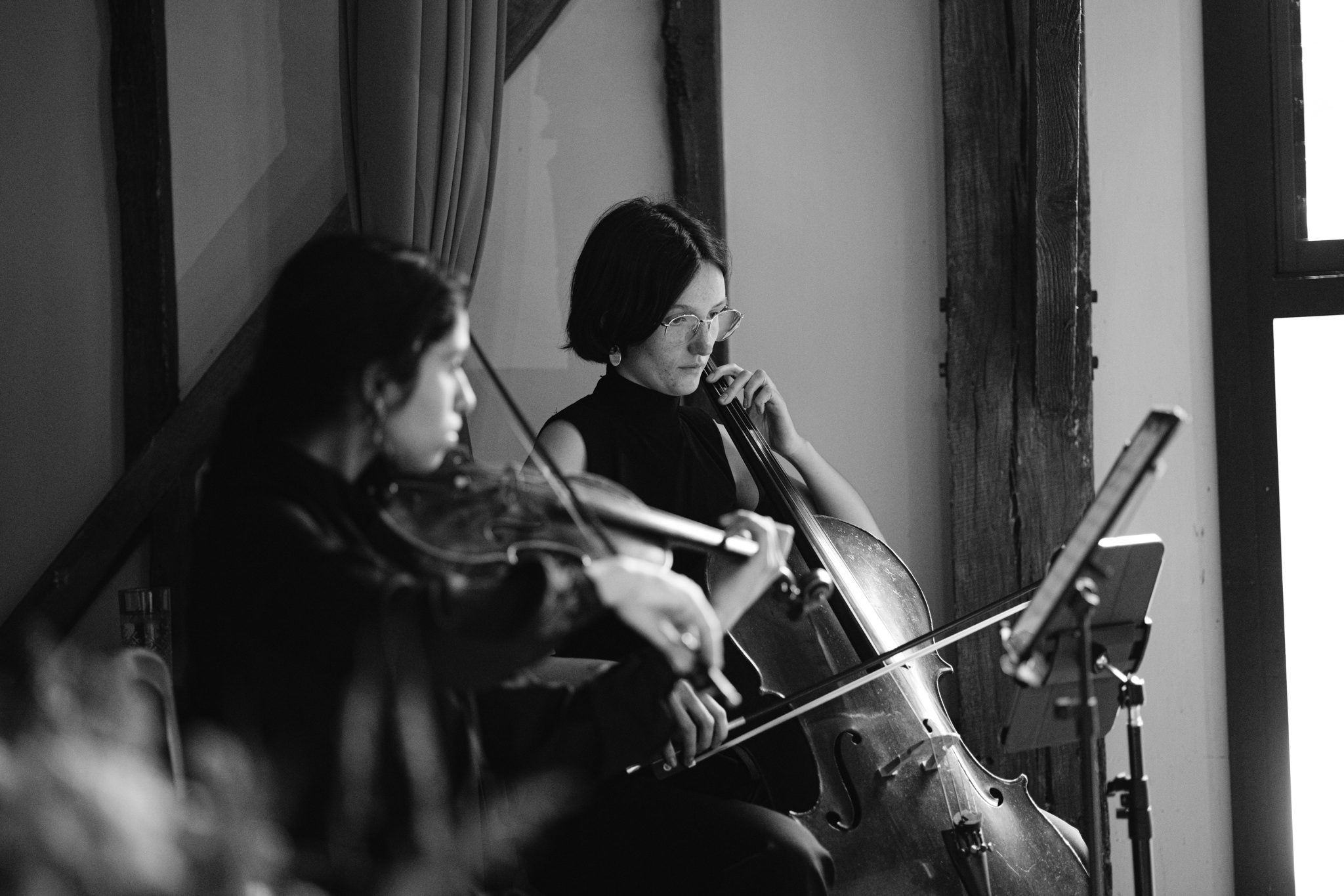 Two musicians perform indoors at a Barn Wedding in Hertfordshire; one plays the violin and the other plays the cello, both reading sheet music from a stand.