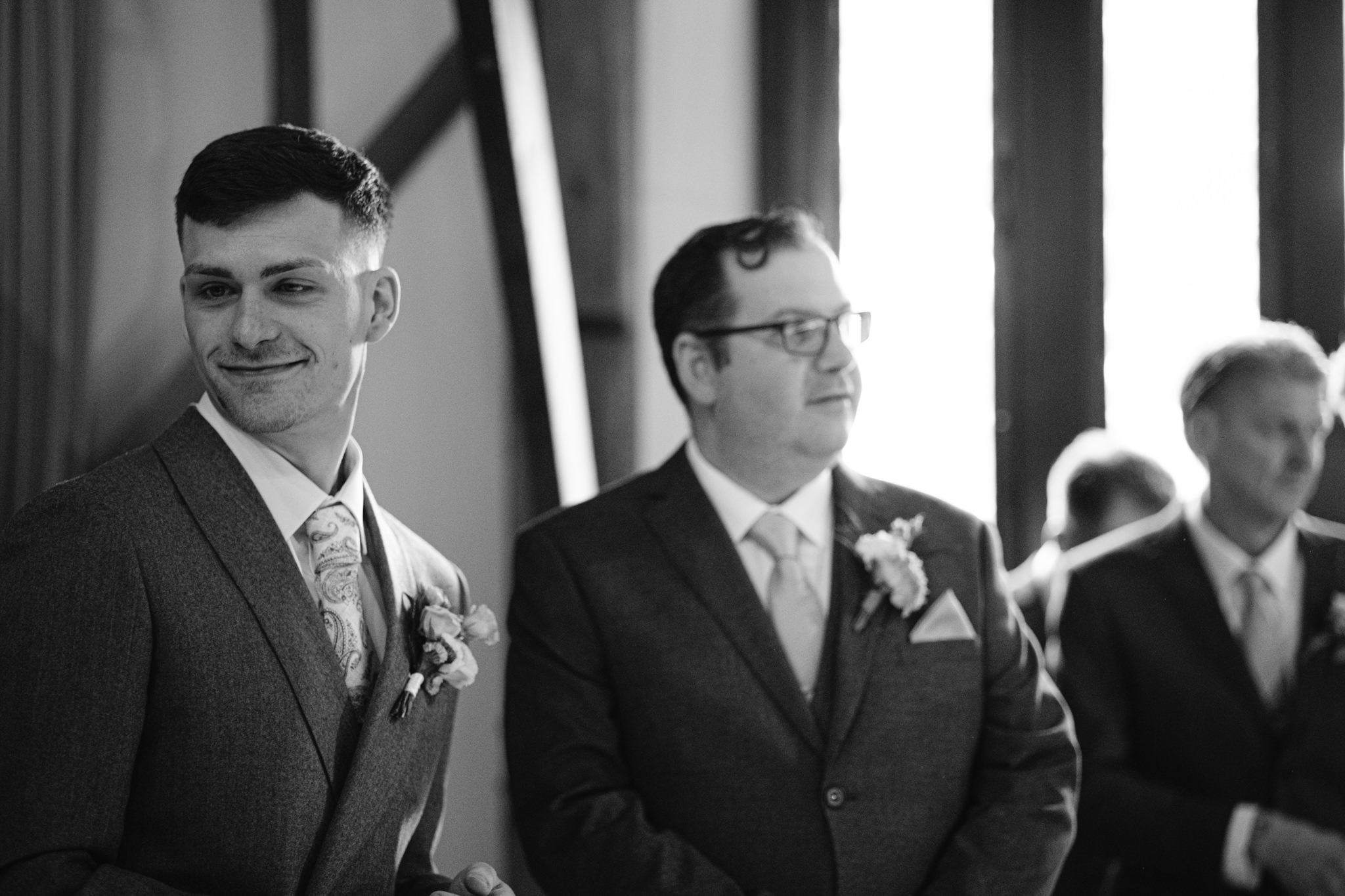 Two men in suits stand indoors at a formal event, with one smiling and the other looking to the side; another man is blurred in the background—capturing candid moments typical of Essendon Country Club Wedding Photography.
