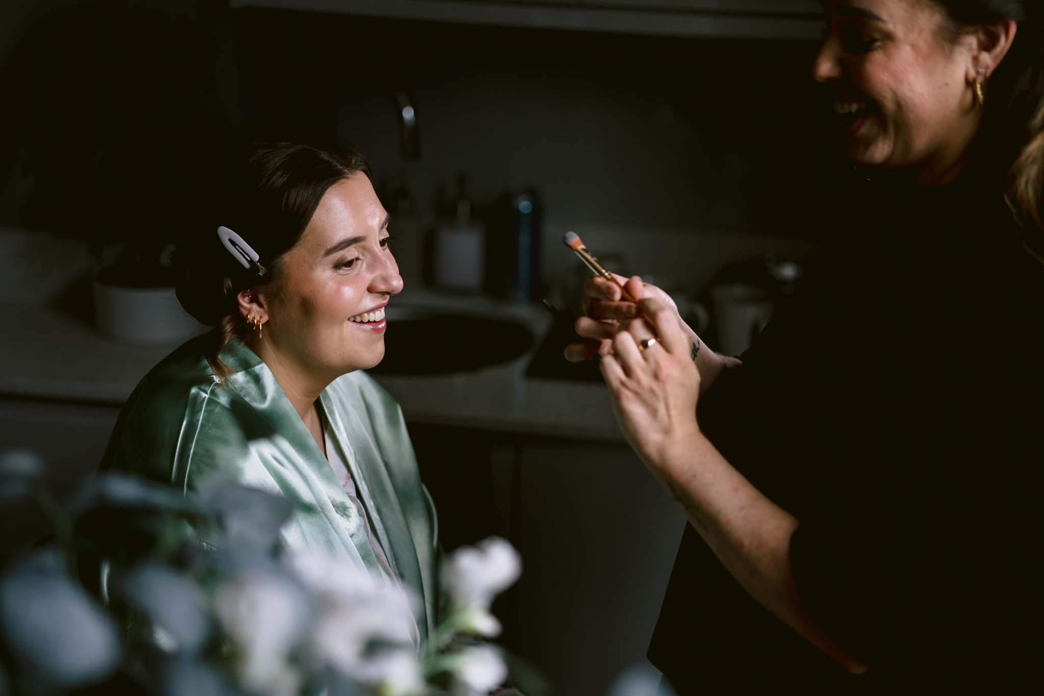 A woman in a green dressing gown sits and smiles whilst another person holds a make-up brush, preparing to apply make-up in the dimly lit room at Essendon Country Club, a beautiful Hertfordshire venue perfect for wedding photography.