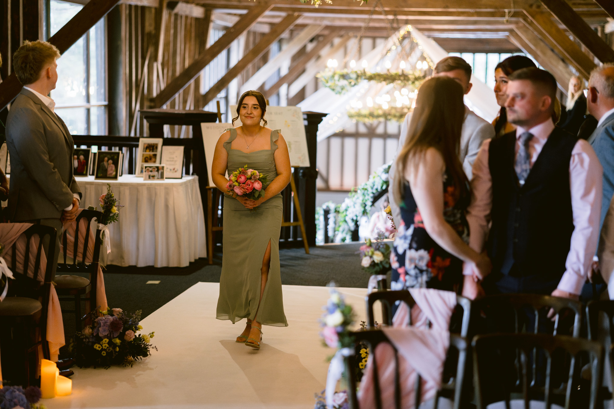 A bridesmaid in a green dress walks down the aisle holding a bouquet, whilst seated guests watch in a rustic, decorated indoor venue—perfect for beautiful Essendon Country Club Wedding Photography.