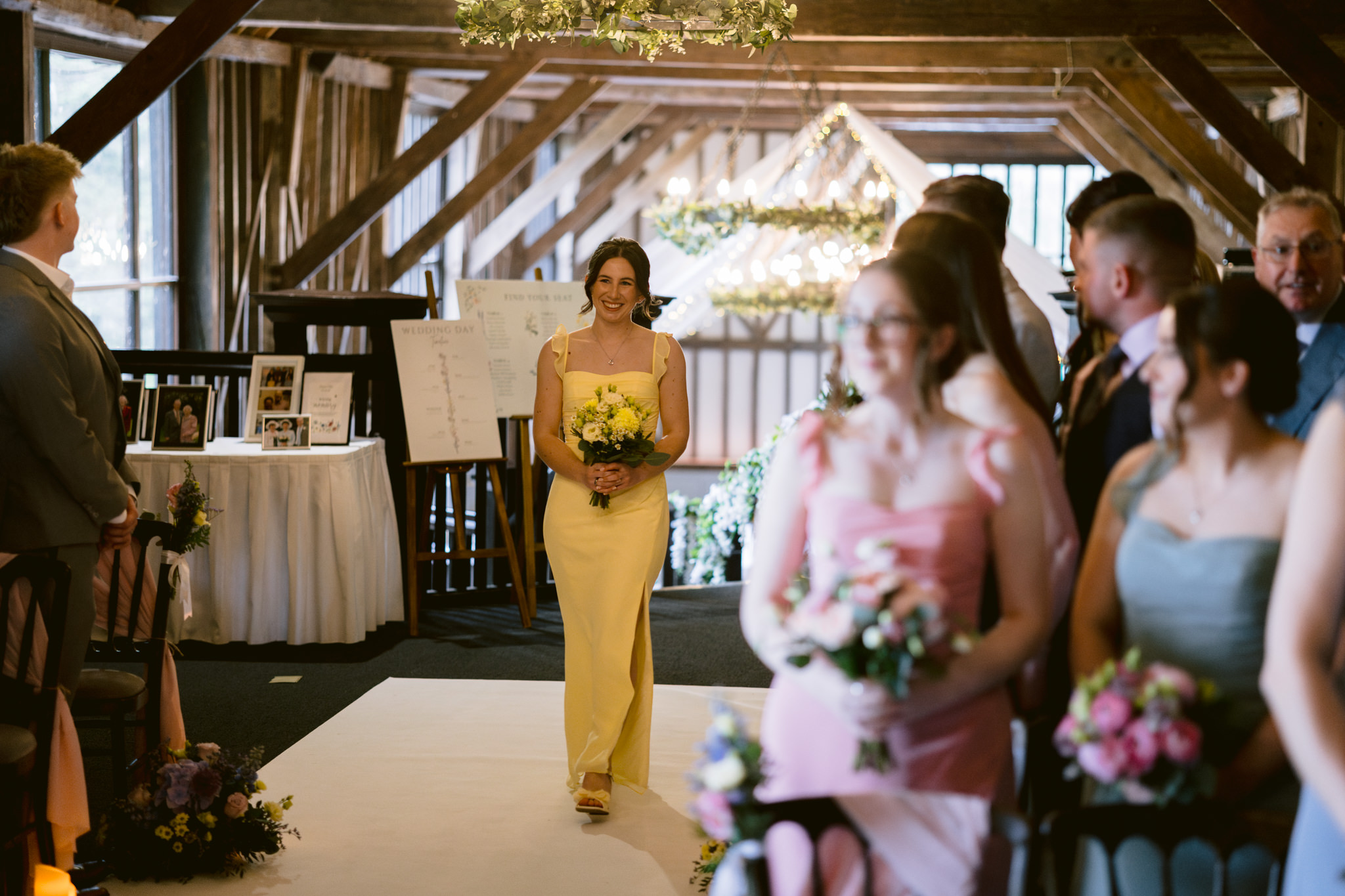 A woman in a yellow dress walks down the aisle holding a bouquet at an indoor wedding ceremony at Essendon Country Club in Hertfordshire, whilst seated guests watch—captured beautifully with expert wedding photography.