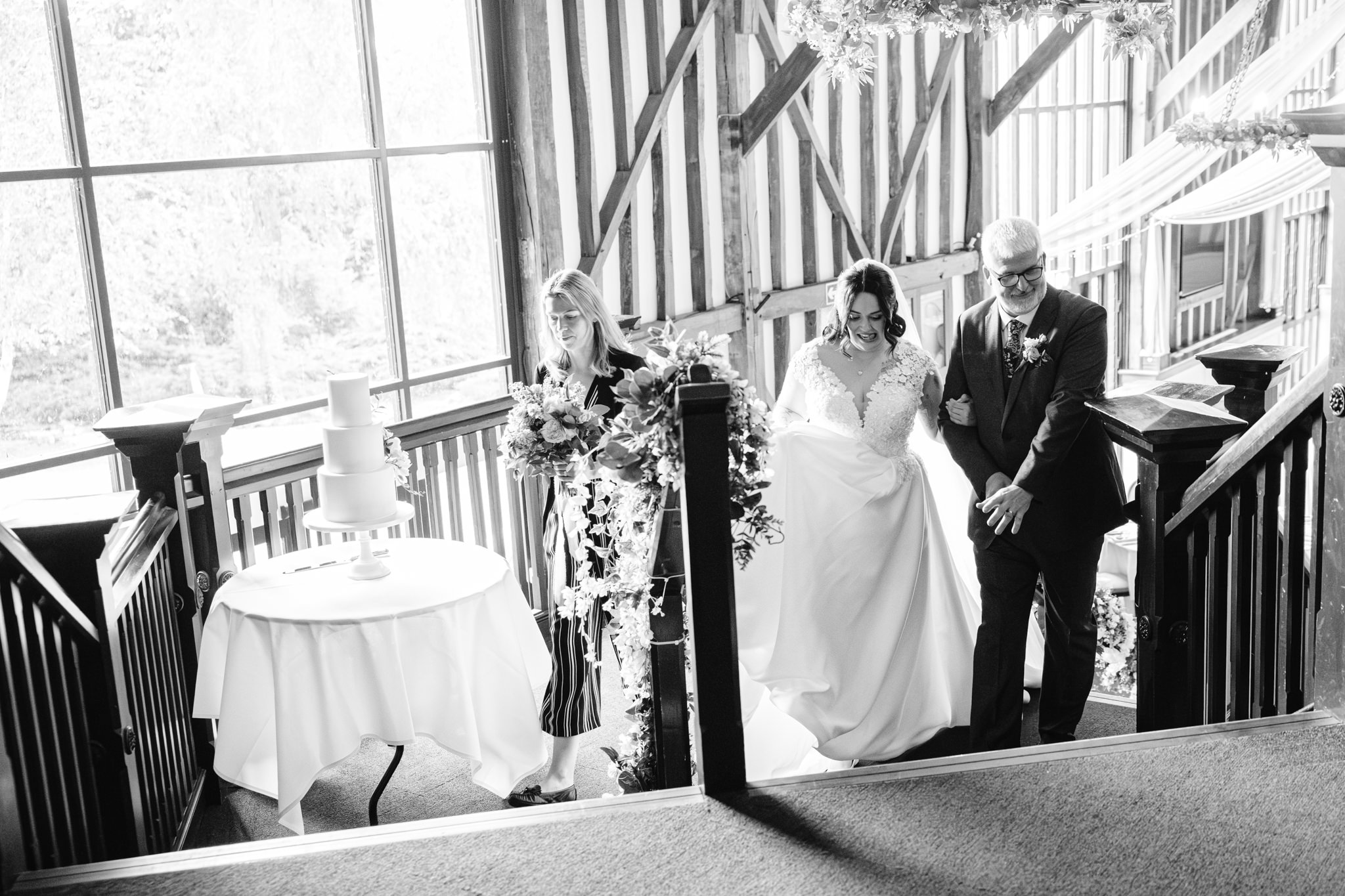 A bride in a white gown walks up stairs with two people; a decorated cake sits on a table by a large window in this charming Essendon Country Club Wedding barn venue.