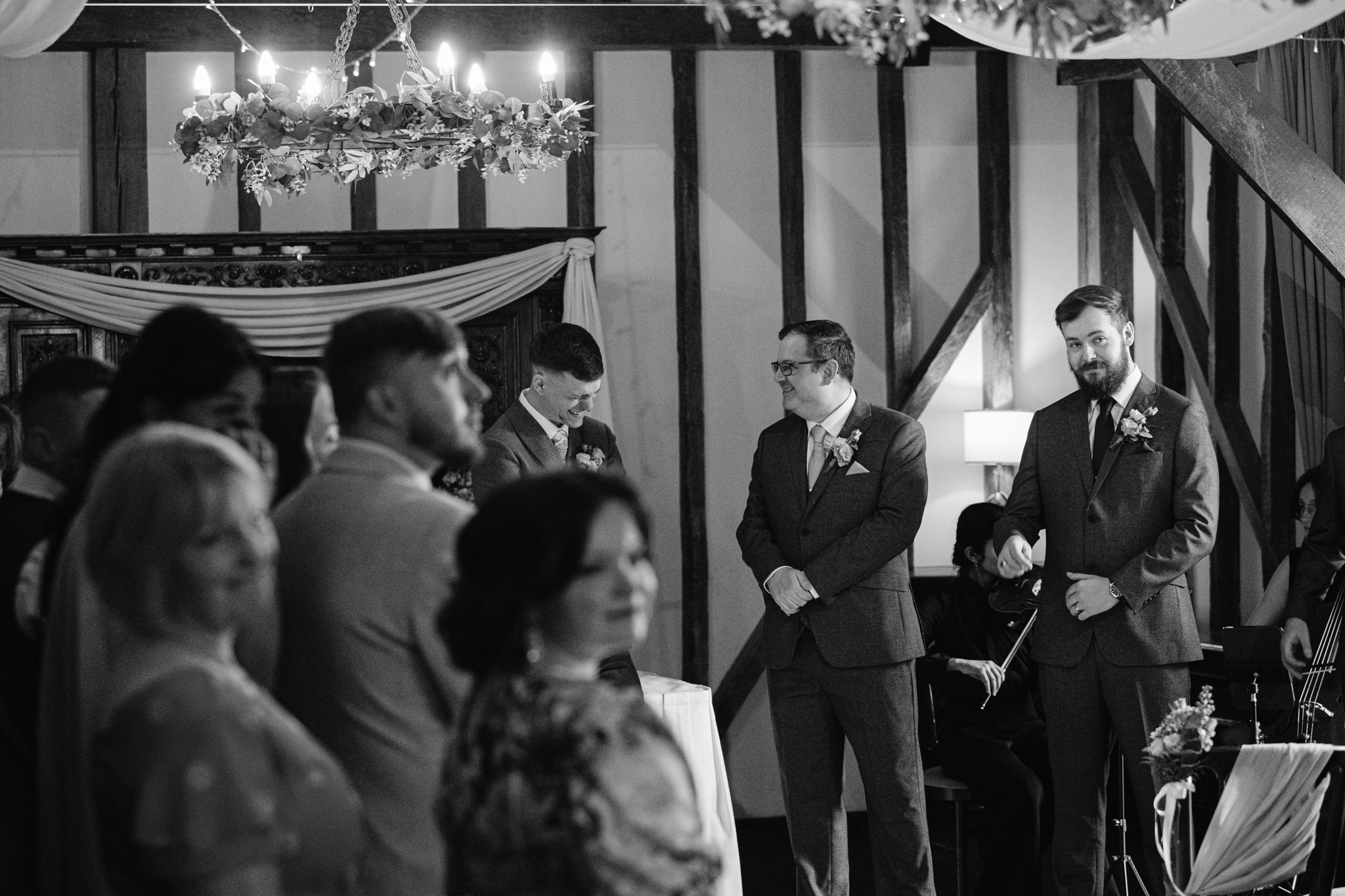 A group of people stand indoors at a Hertfordshire wedding ceremony; two men in suits near the front smile whilst guests look on, capturing a special moment of Essendon Country Club wedding photography.