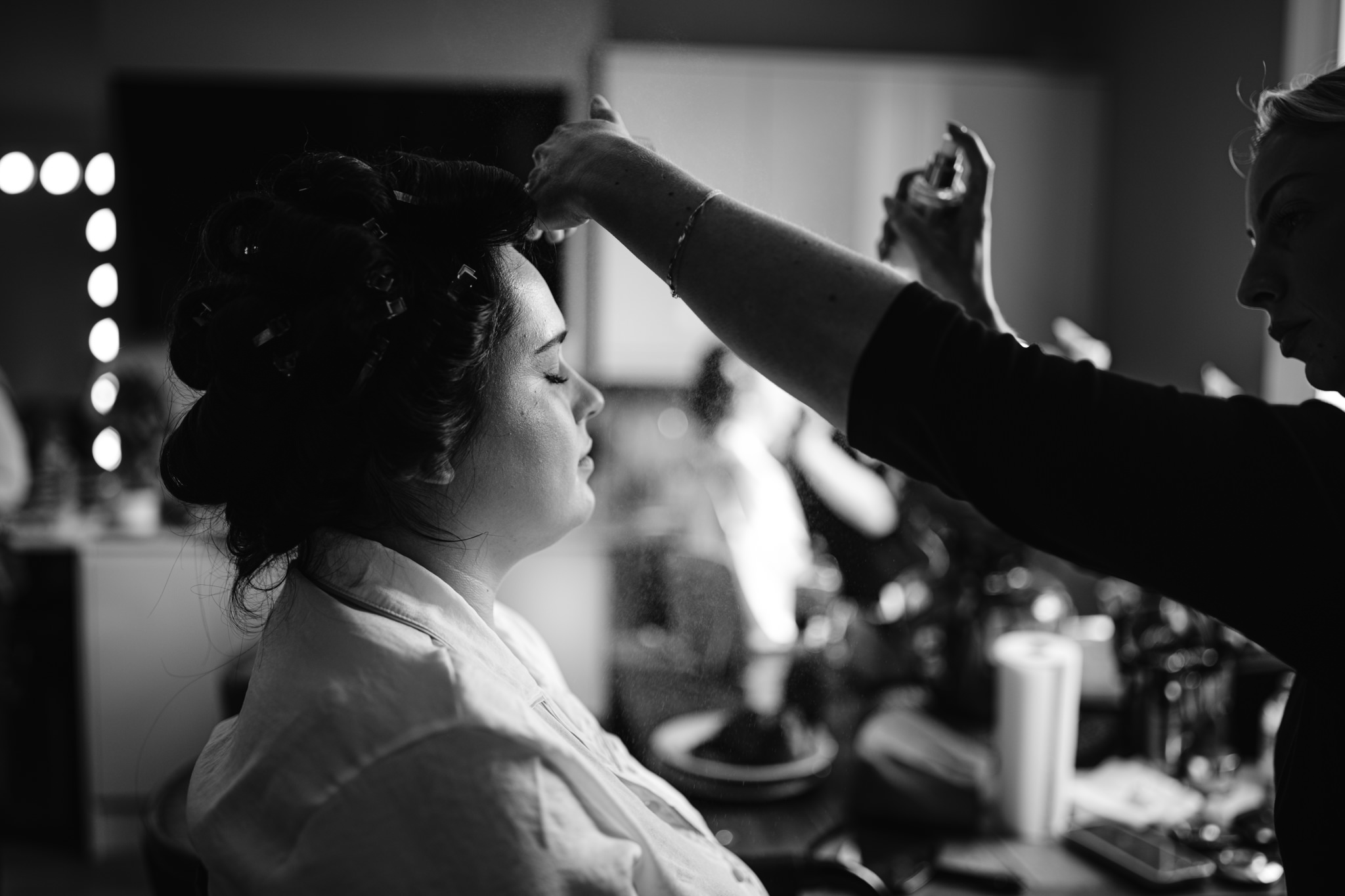 A woman sits with her eyes closed while another person sprays her hair, preparing her in a well-lit room with various objects visible in the background at Essendon Country Club, Hertfordshire—perfect for wedding photography moments.