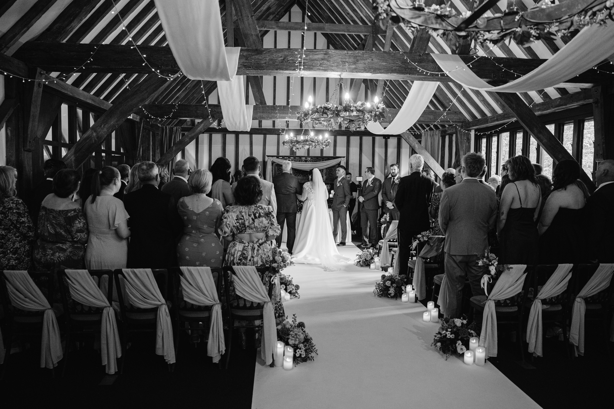 A bride and groom stand at the altar of Essendon Country Club’s decorated wooden hall in Hertfordshire, surrounded by guests during a wedding ceremony—captured in timeless Wedding Photography.