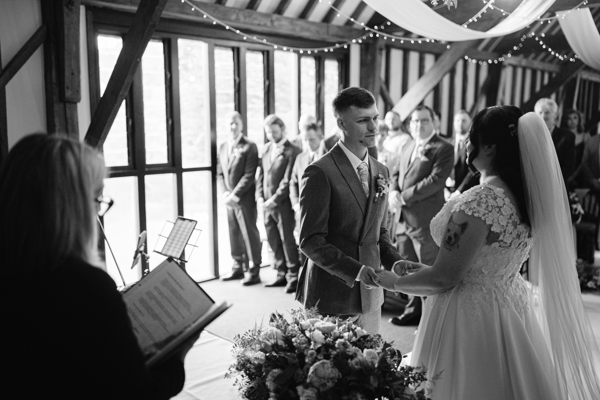 A bride and groom hold hands and face each other during their wedding ceremony in a rustic indoor venue at Essendon Country Club, with guests and an officiant present. Perfect for Barn Wedding Hertfordshire photography moments.