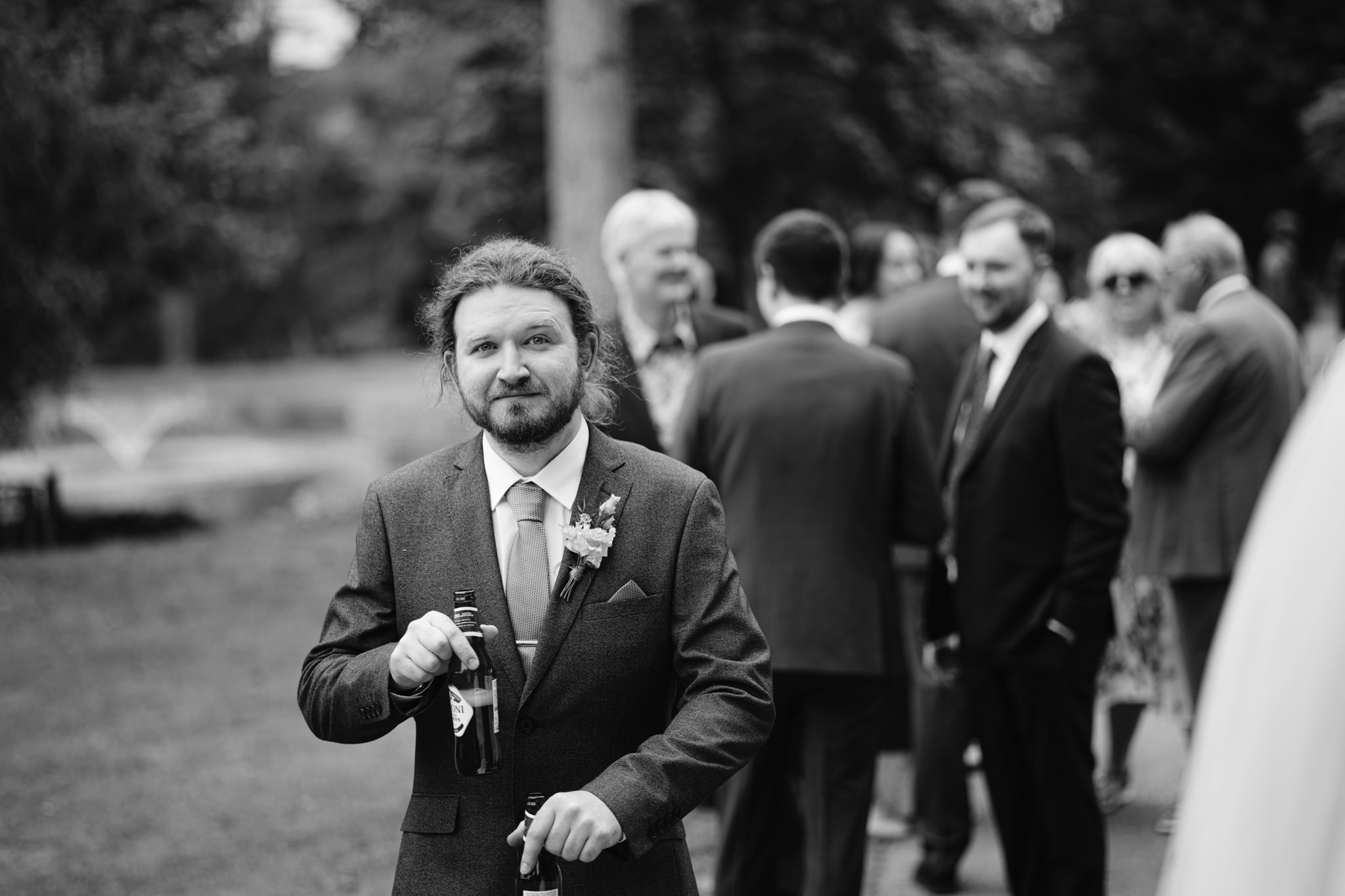 A man in a suit holding two bottles stands outdoors at an Essendon Country Club wedding, with a group of people in formal attire talking in the background.