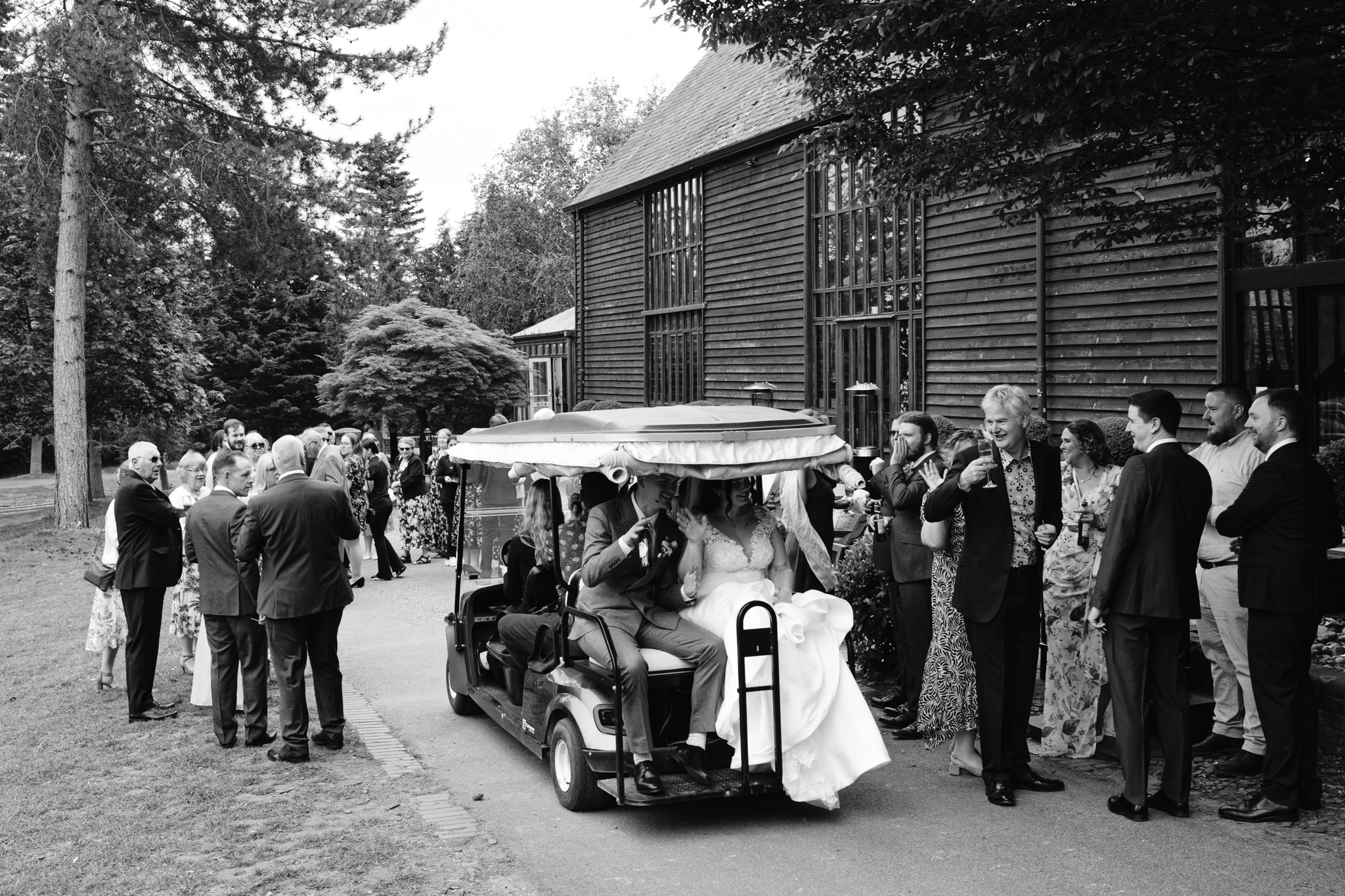 A bride and groom ride a decorated golf buggy past well-dressed guests gathered outside a large wooden building at Essendon Country Club, capturing a joyful moment of their Hertfordshire wedding photography.