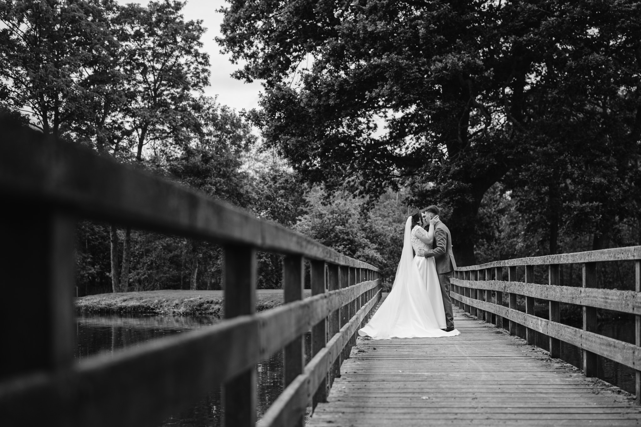 A bride and groom share a kiss on a wooden bridge surrounded by trees in this black and white Essendon Country Club Wedding Photography outdoor photo.