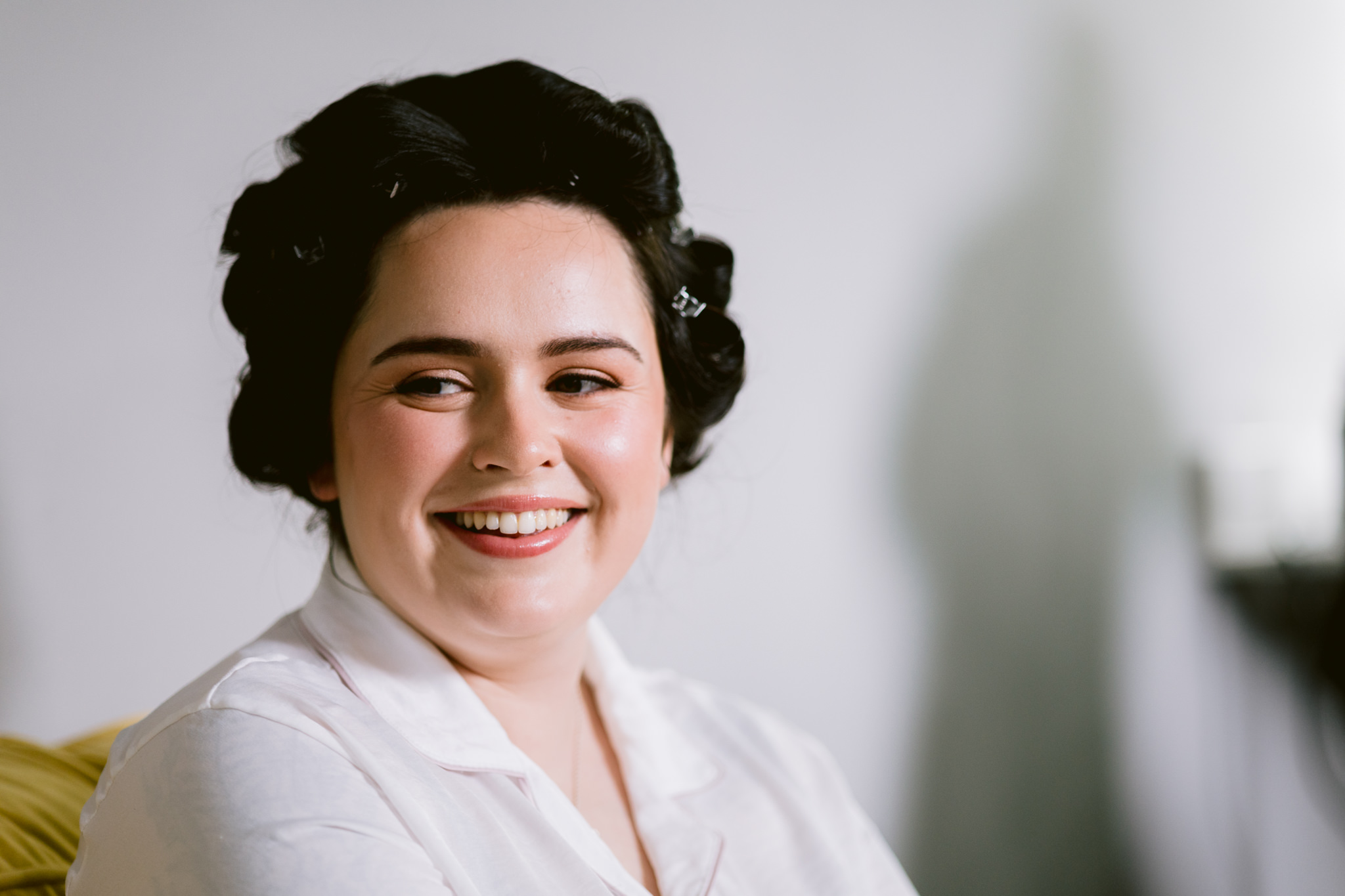 A person with dark hair in rollers smiles whilst wearing a white shirt, seated indoors with a blurred background—capturing the joyful preparation before a Barn Wedding Hertfordshire.