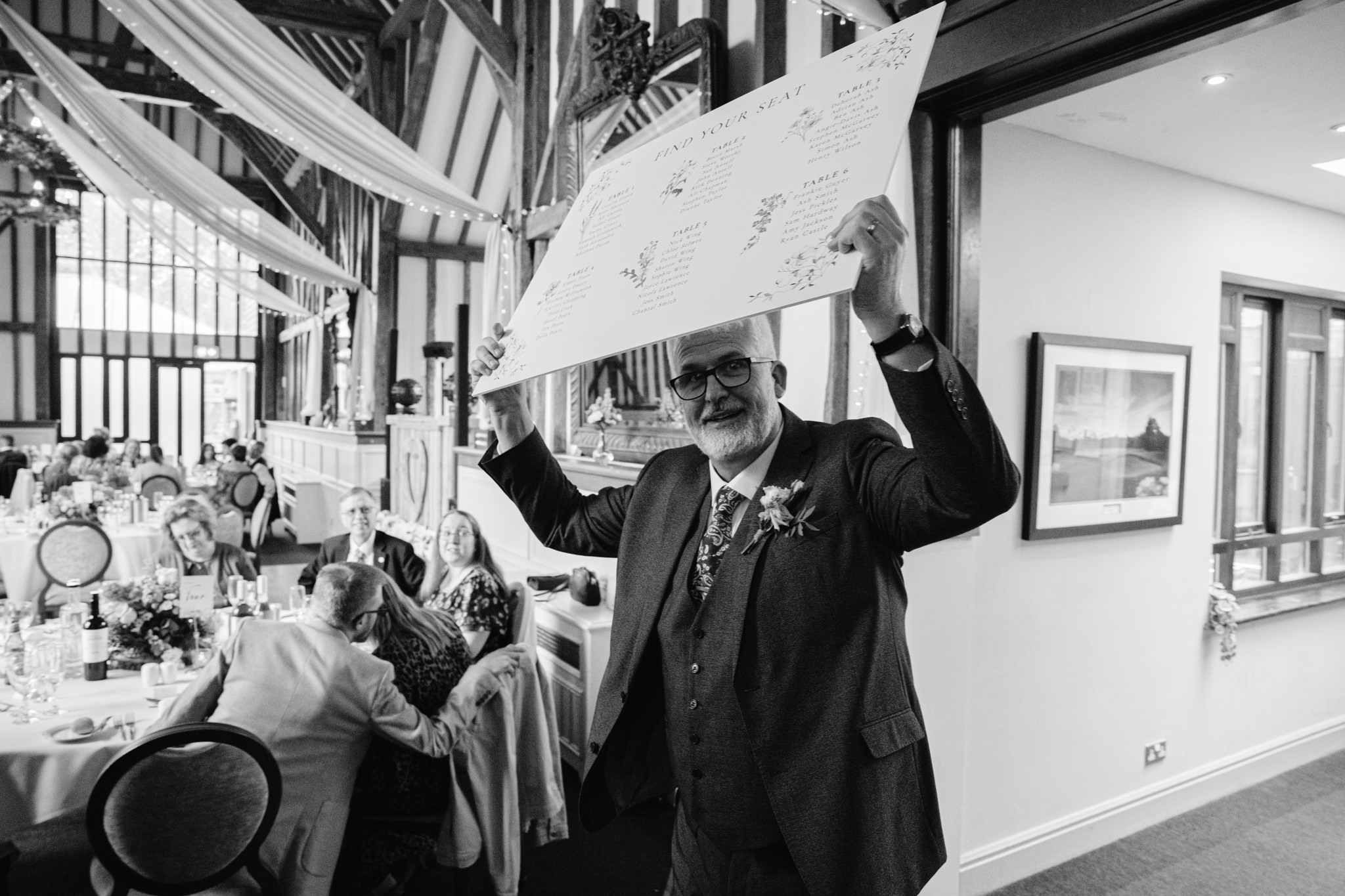 A man in a suit holds up a large seating plan at a formal Essendon Country Club wedding, with guests seated at decorated tables—an elegant moment perfect for wedding photography.