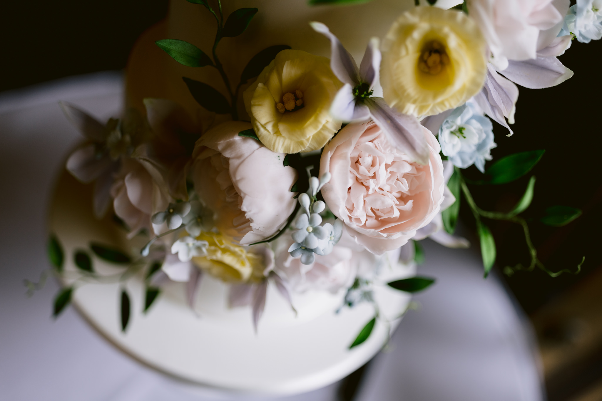 Close-up of a white cake decorated with pastel pink and yellow flowers, green leaves, and small white berry-like accents—perfect for capturing in Hertfordshire wedding photography at Essendon Country Club.