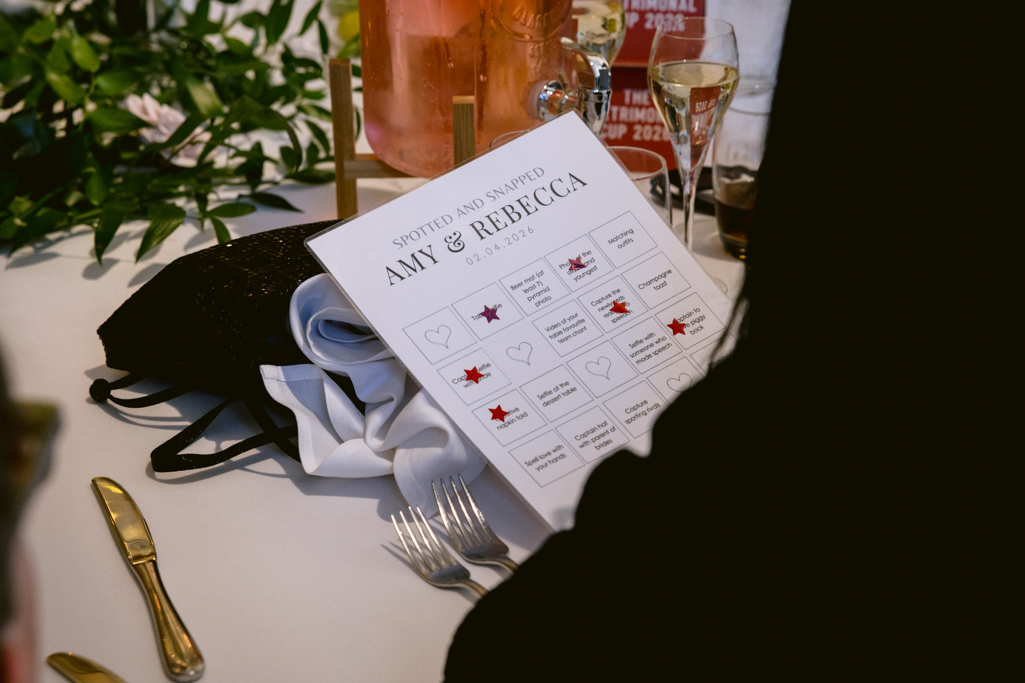 A person holds a "Spotted and Snapped" game card for Amy and Rebecca's event at Upwaltham Barns Wedding Photography, placed on a table set with cutlery, ribbon, and drinks.