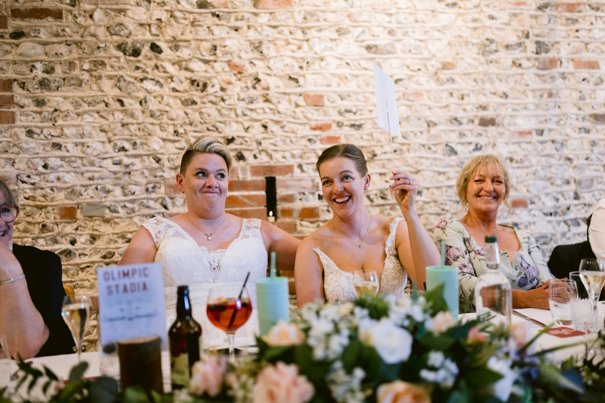 Two women in wedding dresses sit at a decorated table, smiling, as one raises a piece of paper. Other guests are nearby against a brick wall background, capturing the joy of Upwaltham Barns Wedding Photography.