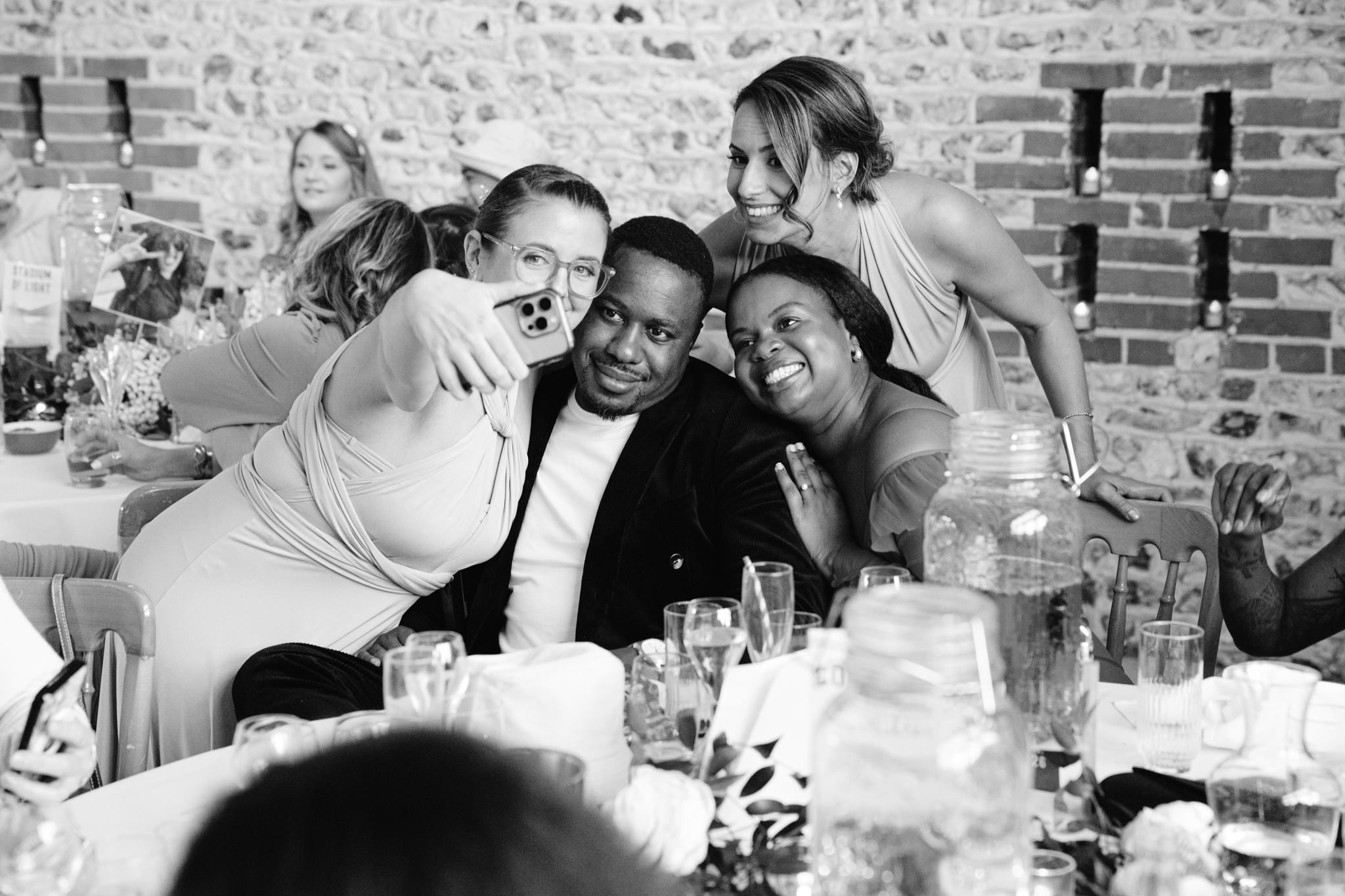 Four people at a formal event sit at a decorated table, smiling and posing for a selfie together, drinks and jars visible in the foreground—a perfect moment captured in an Upwaltham Barns Wedding Photography style.