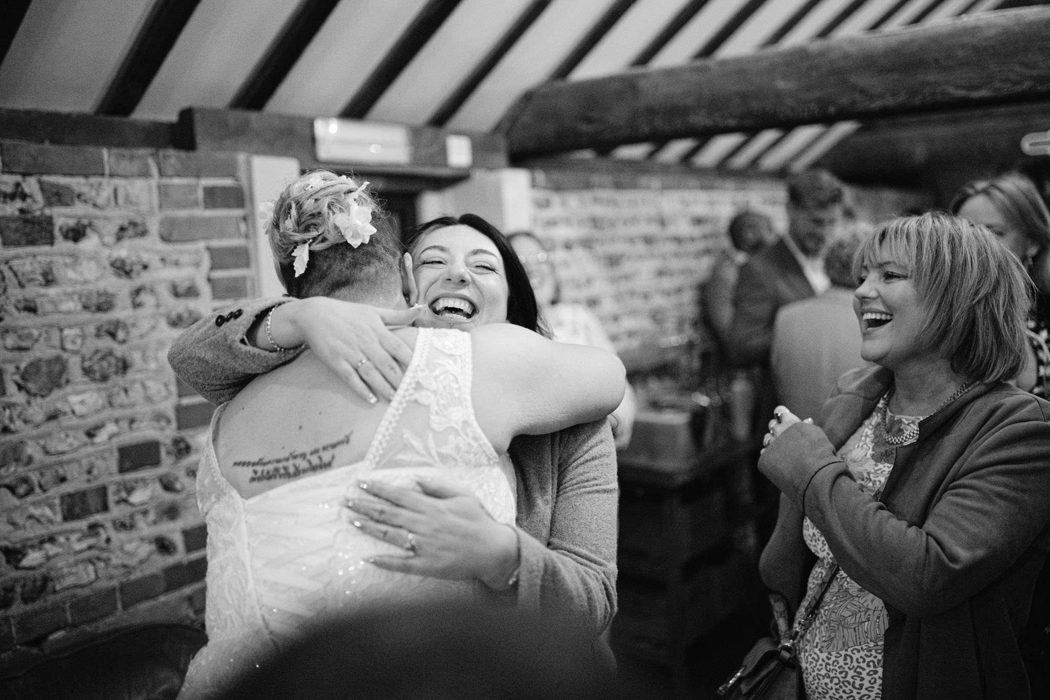 A bride in a white dress hugs a smiling woman whilst another laughs nearby, all set against brick walls—capturing the joy of an Upwaltham Barns Wedding Photography moment indoors.