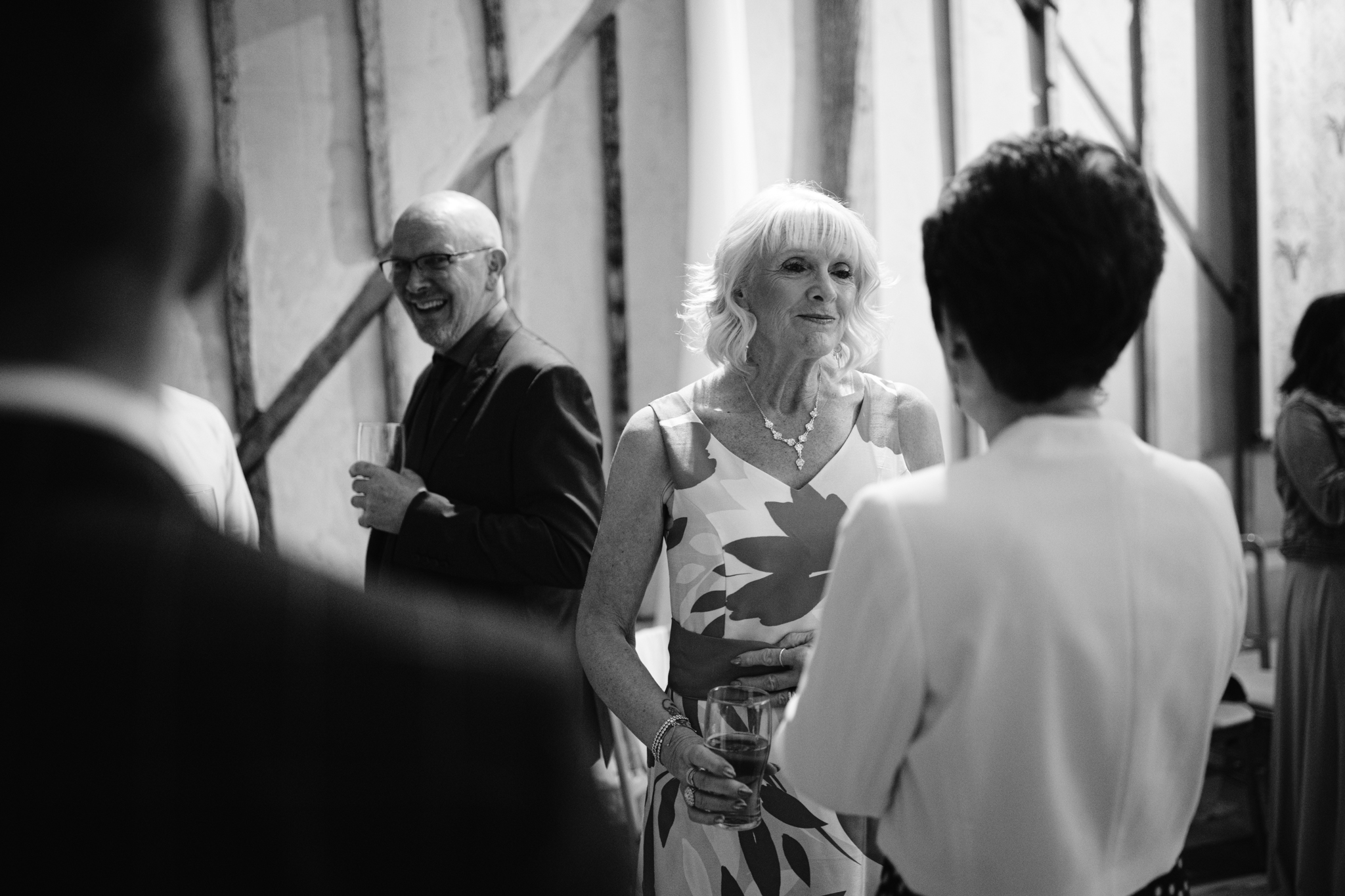 A woman in a floral dress holds a drink and chats with another guest at a lively social gathering, with other guests mingling in the background—capturing the charm of Upwaltham Barns Wedding Photography.