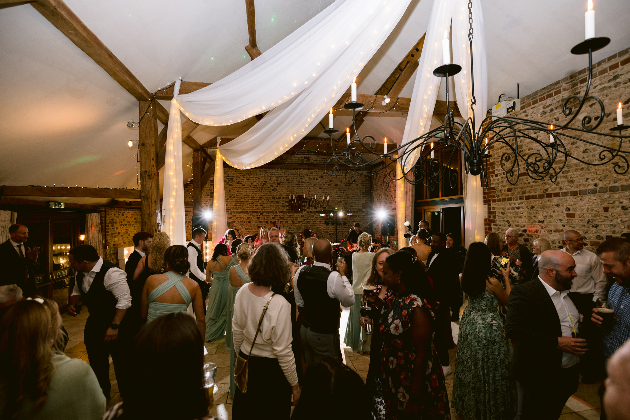 A group of people dressed in formal attire gather and mingle under draped white fabric and chandeliers in a rustic venue with exposed brick walls, perfectly capturing the elegance of Upwaltham Barns Wedding Photography.