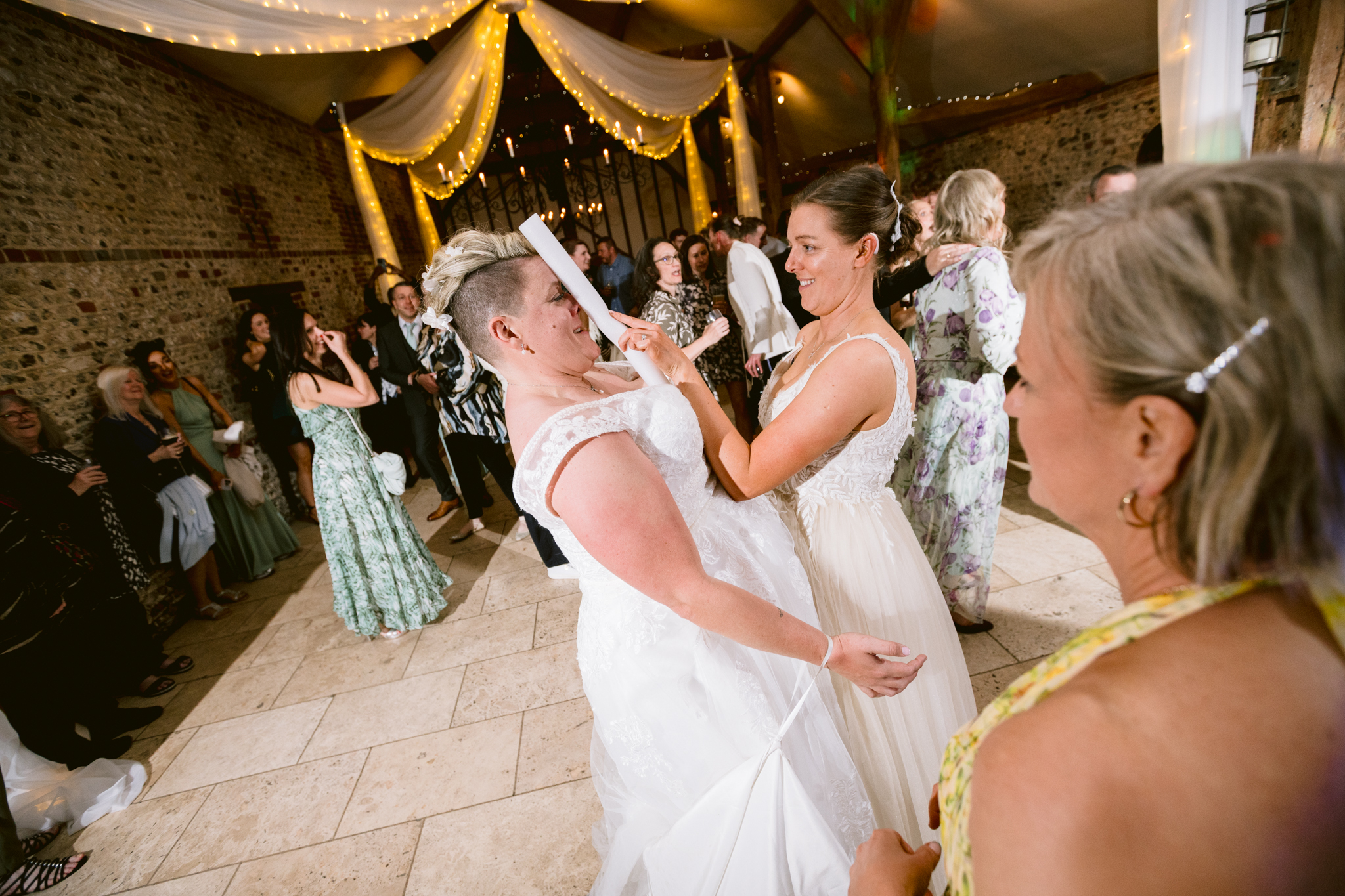 Two brides in wedding dresses laugh and sing whilst holding a rolled-up scroll on a decorated dance floor, surrounded by guests at their joyful Upwaltham Barns Wedding Photography reception.